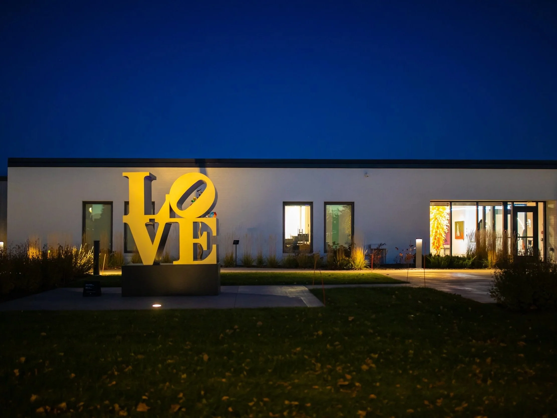 A building with a large illuminated yellow 'LOVE' sign in front of it. The building has a white facade with windows and a well-lit interior. The sky is dark, indicating night time.