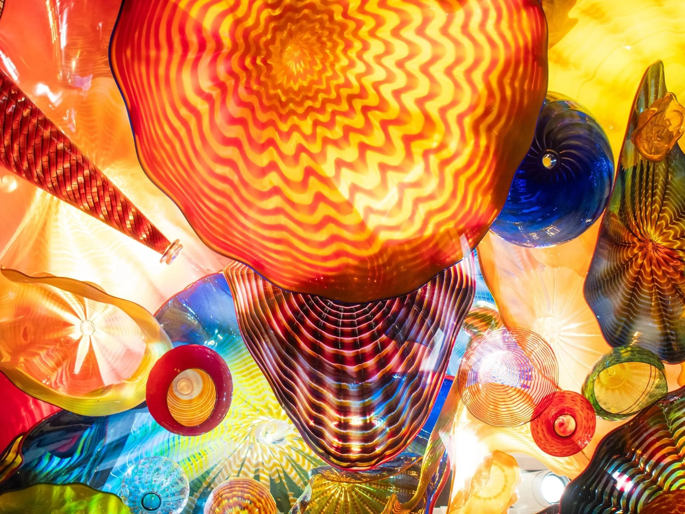 Colorful glass art installation featuring a variety of vibrant, swirling glass pieces in red, yellow, blue, and green with intricate patterns and textures.