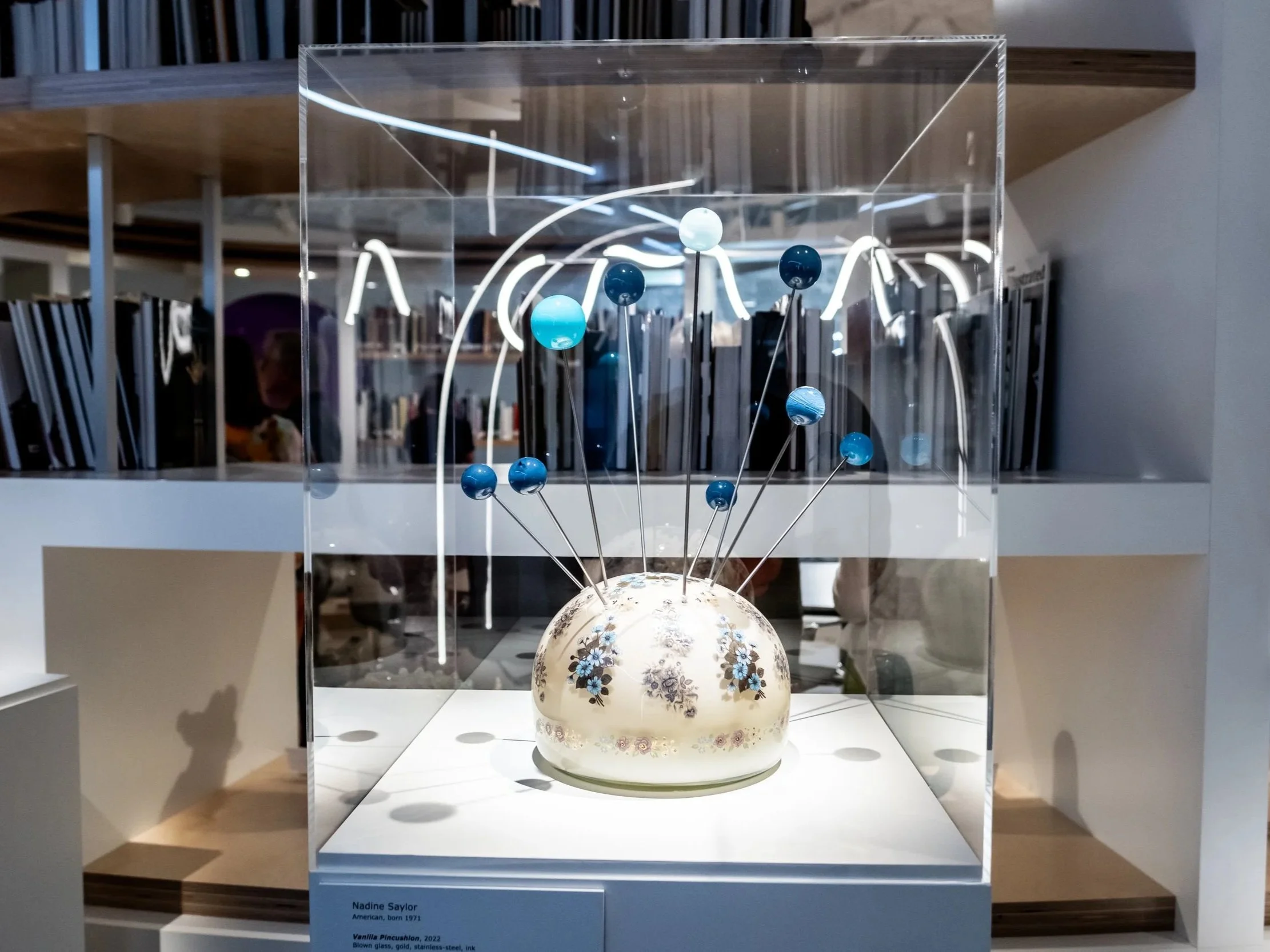 A glass display case contains a ceramic object resembling a small globe with floral designs, with metal rods holding small blue spheres representing planets, and white light lines depicting planetary orbits.