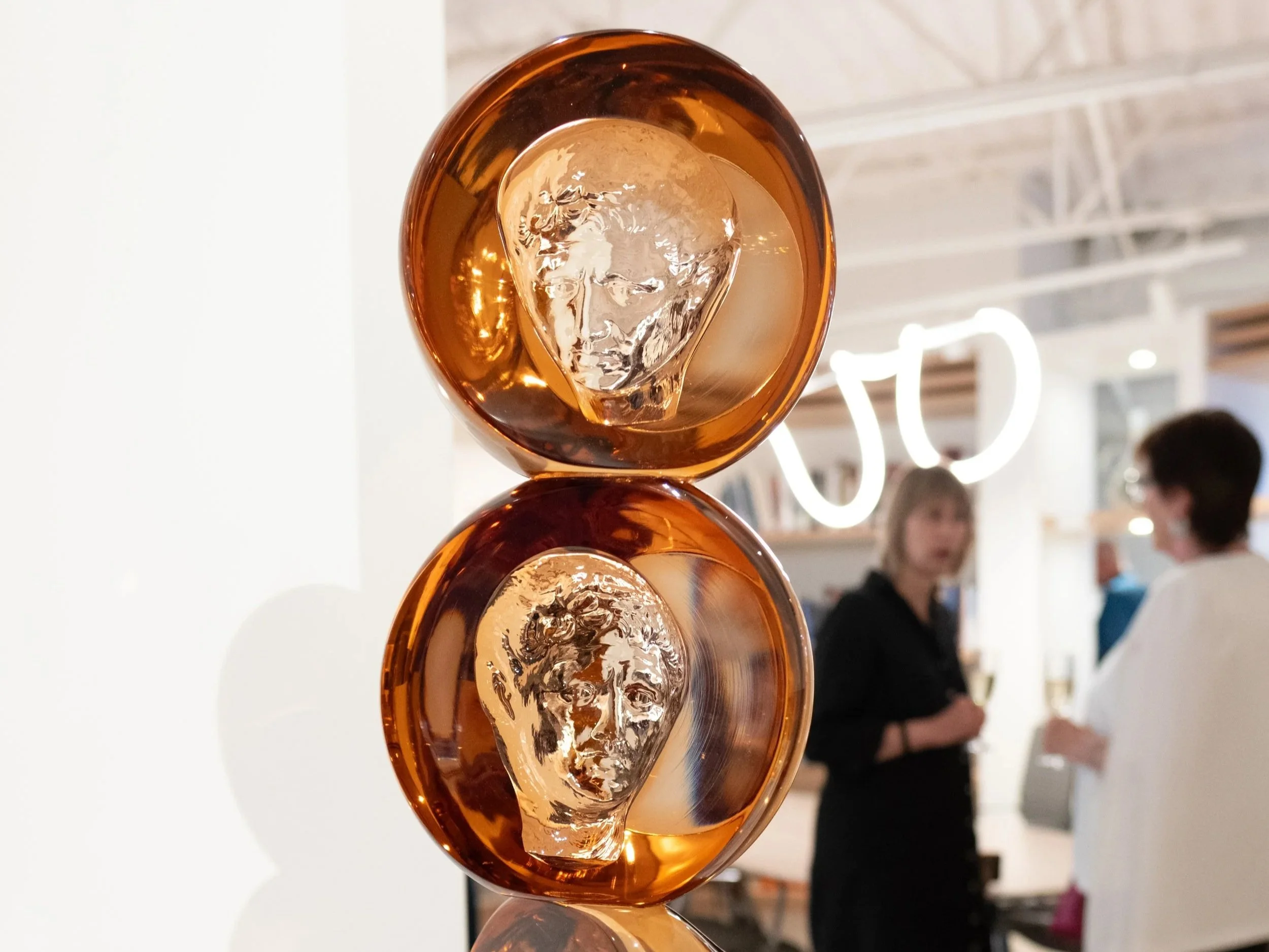 Close-up of three stacked amber-colored glass bowls with textured glass inserts, reflective surfaces, in a modern store or gallery setting with blurred background.