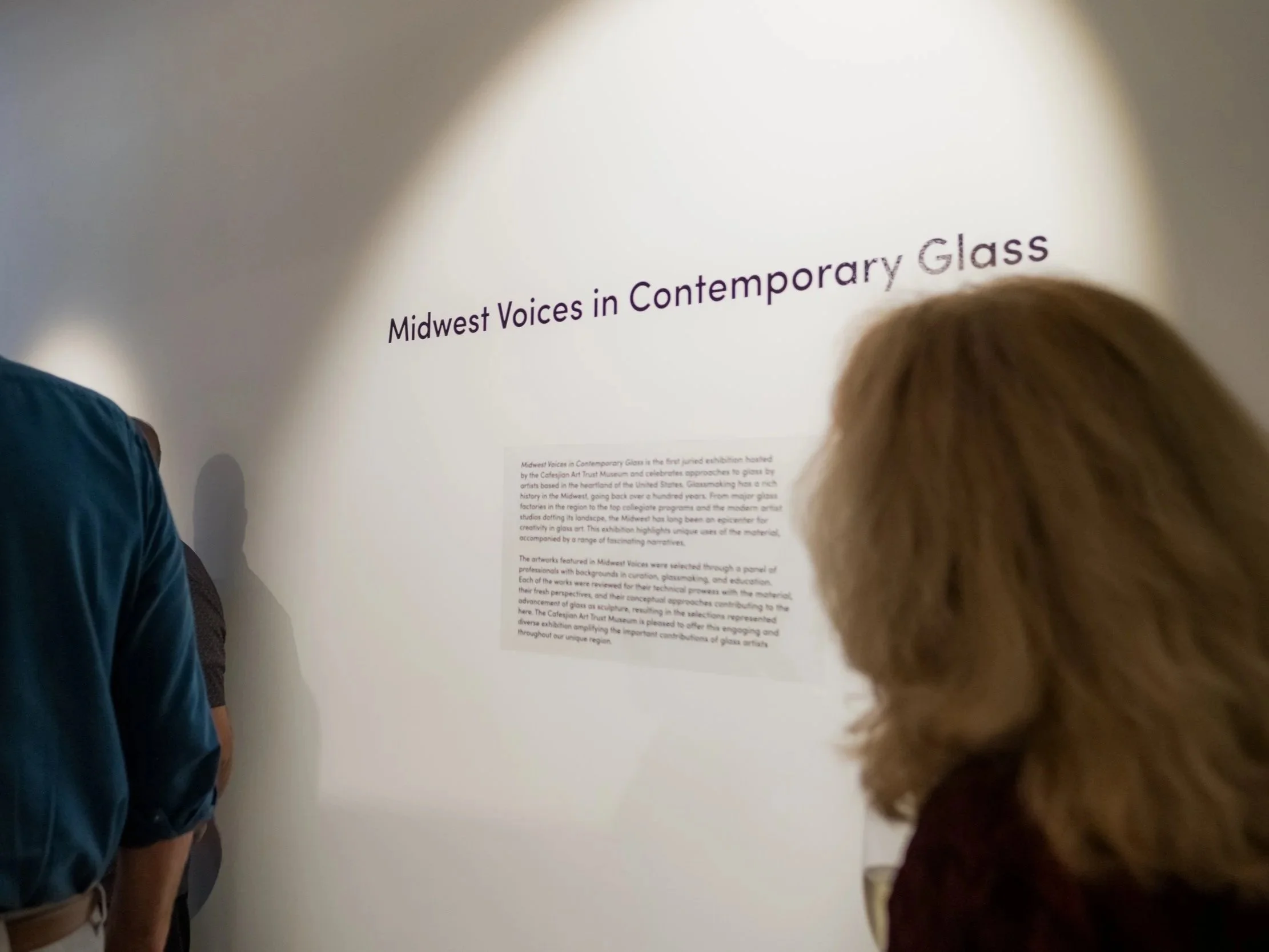 Two people in a museum or gallery looking at an informational display about Midwest Voices in Contemporary Glass.