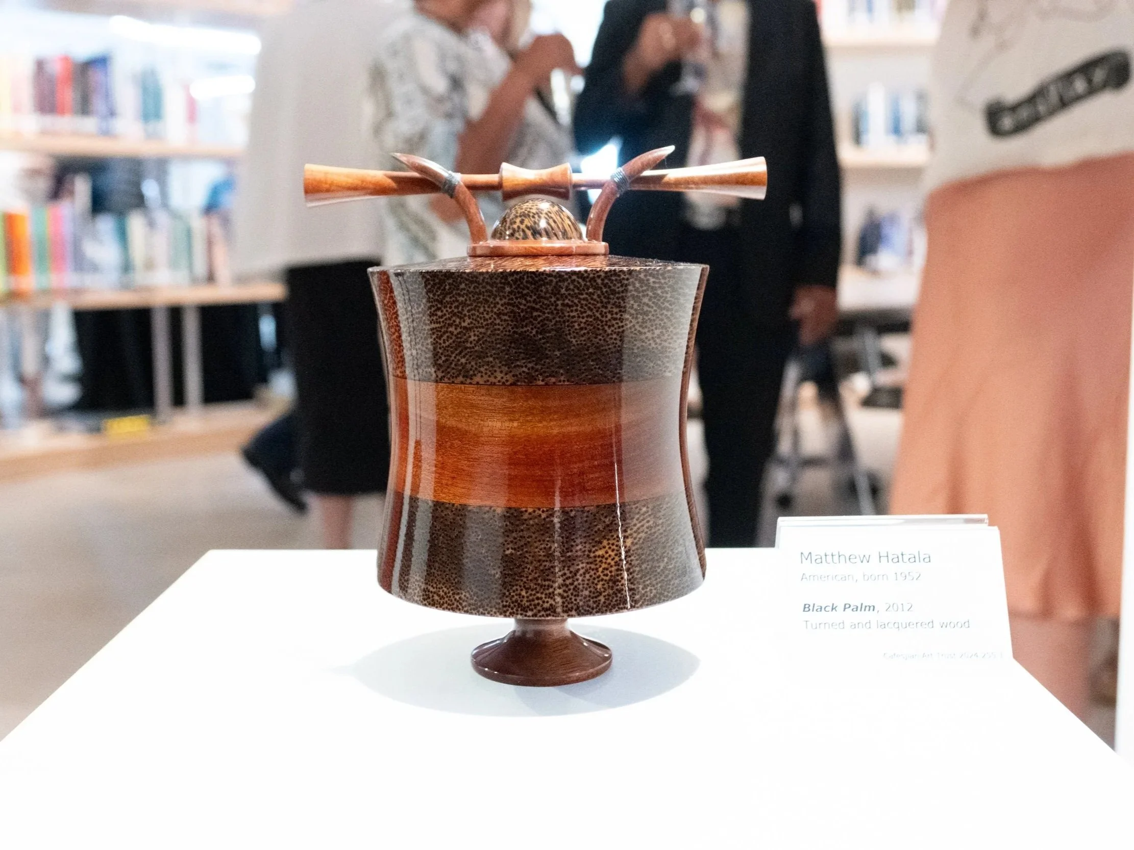 A wooden vase with a narrow base and a wide body, decorated with a black palm motif, displayed on a white surface at an exhibition. There are people and shelves with books in the blurred background.