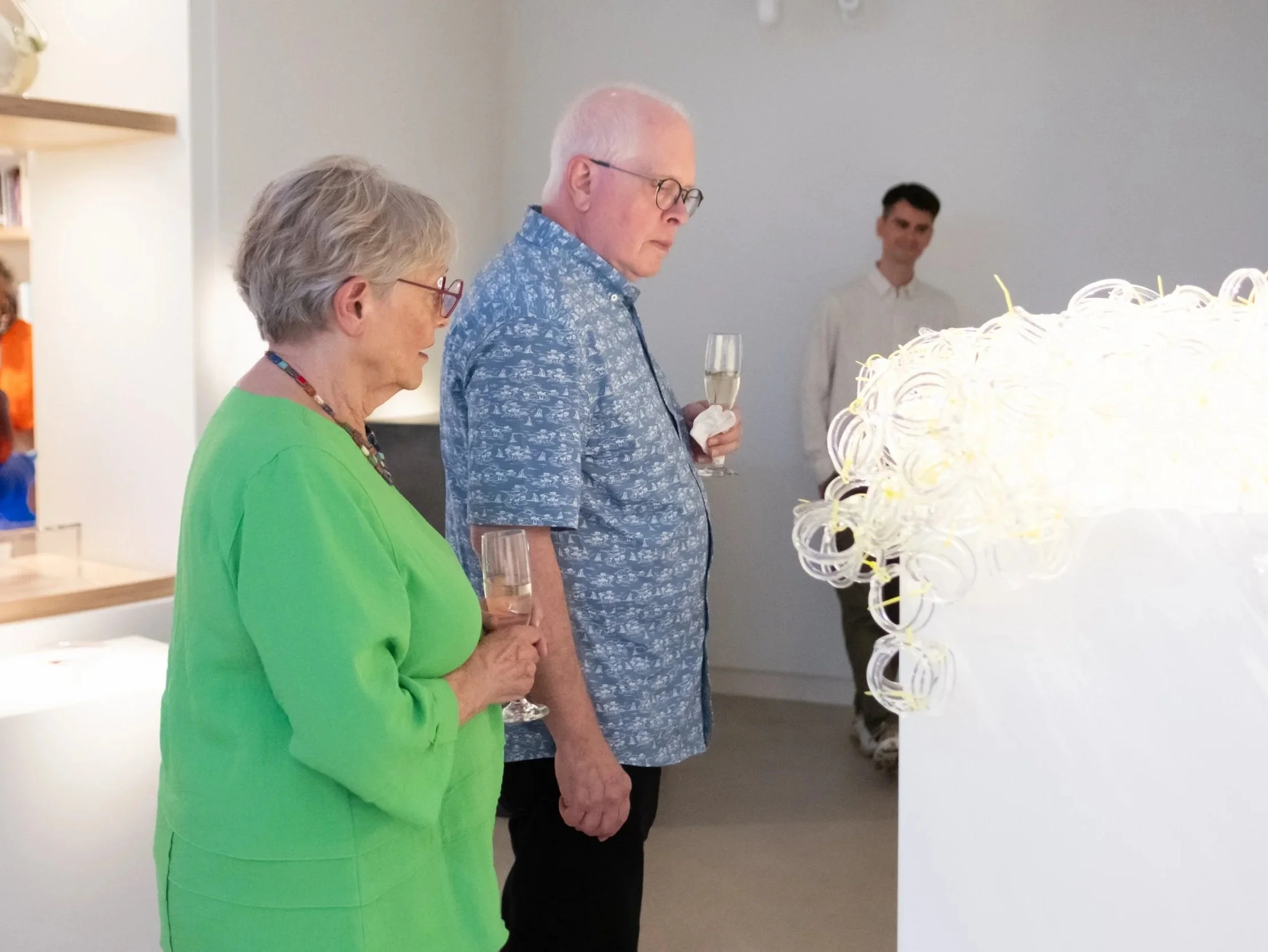 Three people are standing and observing a large white sculpture made of intricate coiled loops. The sculpture appears to be modern art. The individuals are dressed casually and are inside an art gallery or exhibition space.