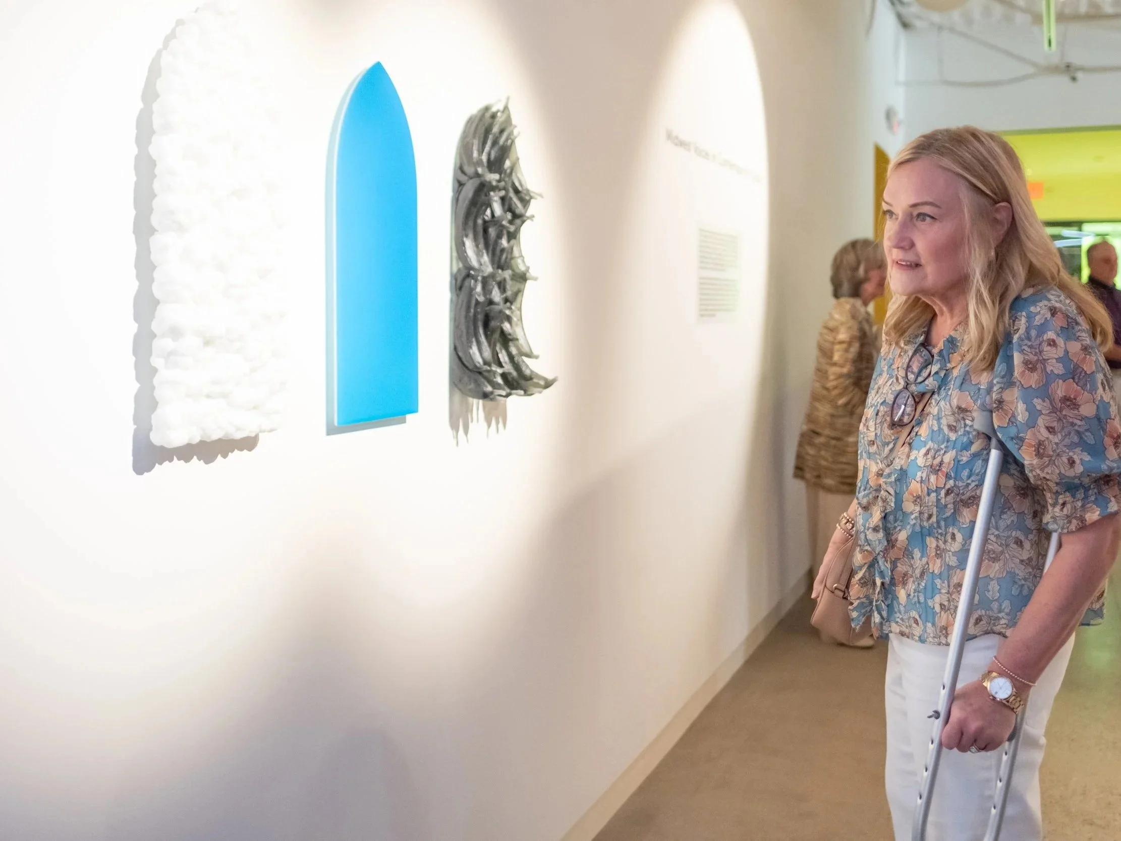 A woman wearing a floral blouse and white pants looking at artwork in a museum, using a cane, with other visitors in the background.