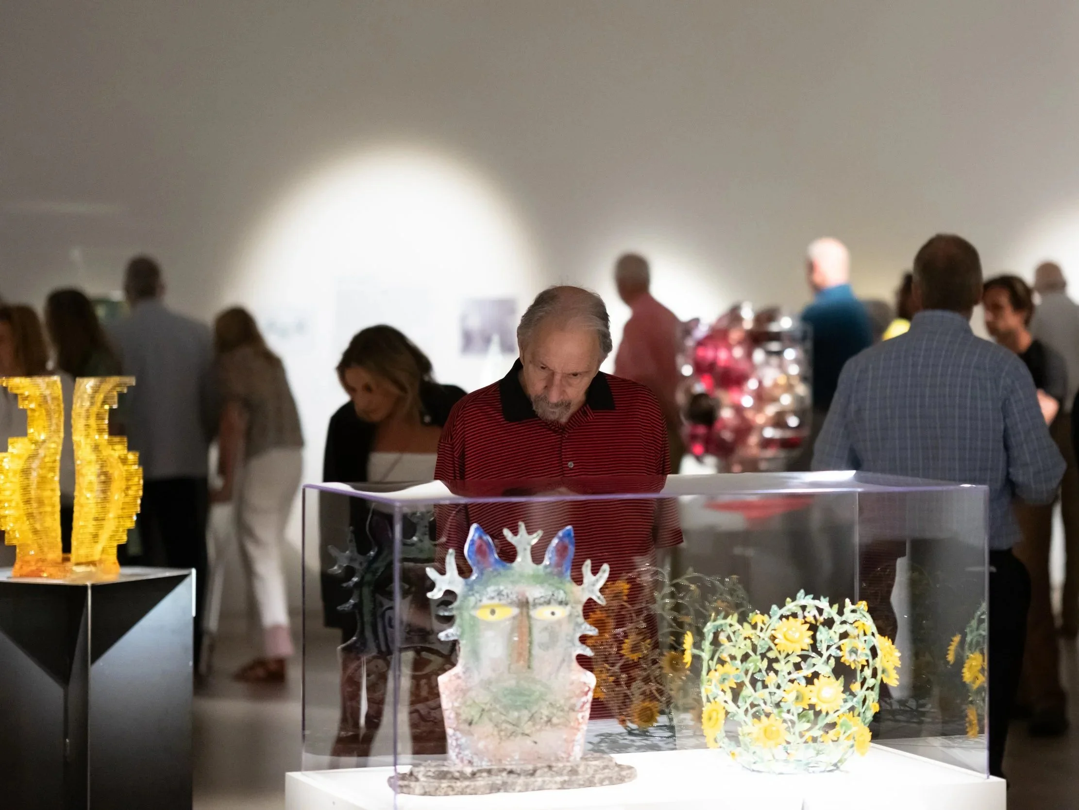 A man with long gray hair and a beard wearing a red and black striped polo shirt is looking down at a display case with colorful ceramic or decorated glass art pieces. The background shows a group of people gathered around, illuminated by soft overhe