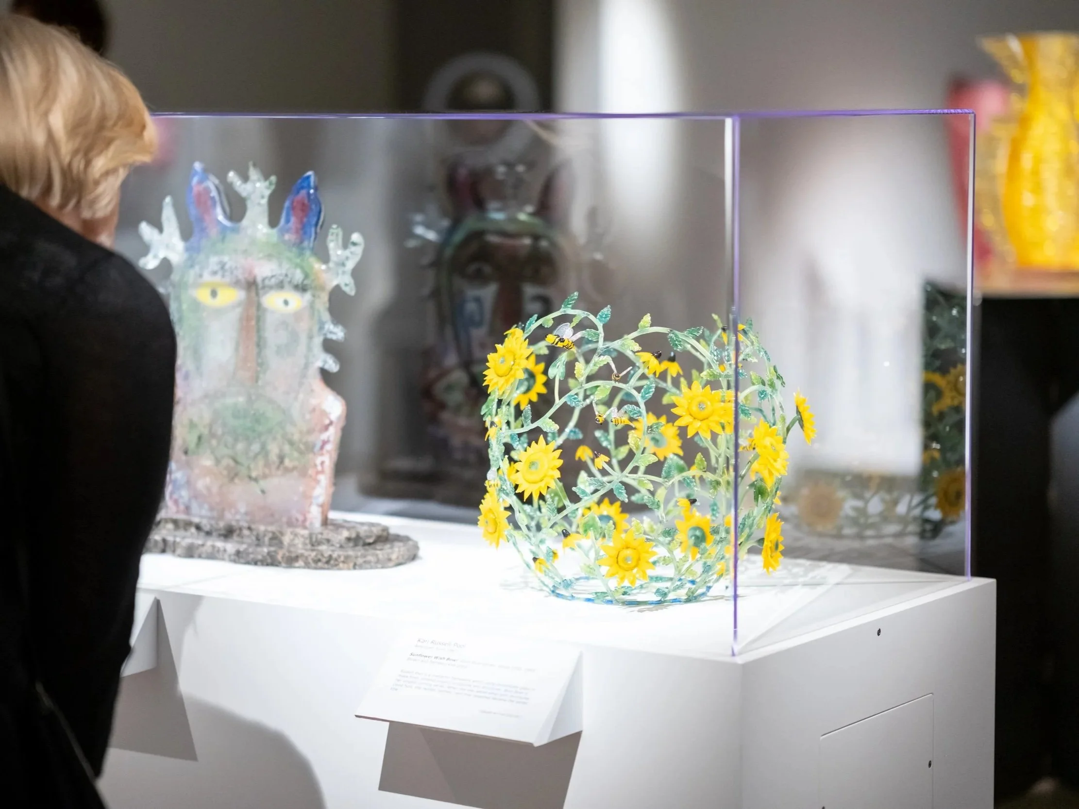 A colorful glass sculpture of a face with rainbow-colored hair and a vibrant, elaborate crown of snowflakes. Next to it, a detailed glass sunflower and leaves arrangement in a clear glass bowl is displayed inside a glass case at an art exhibit.