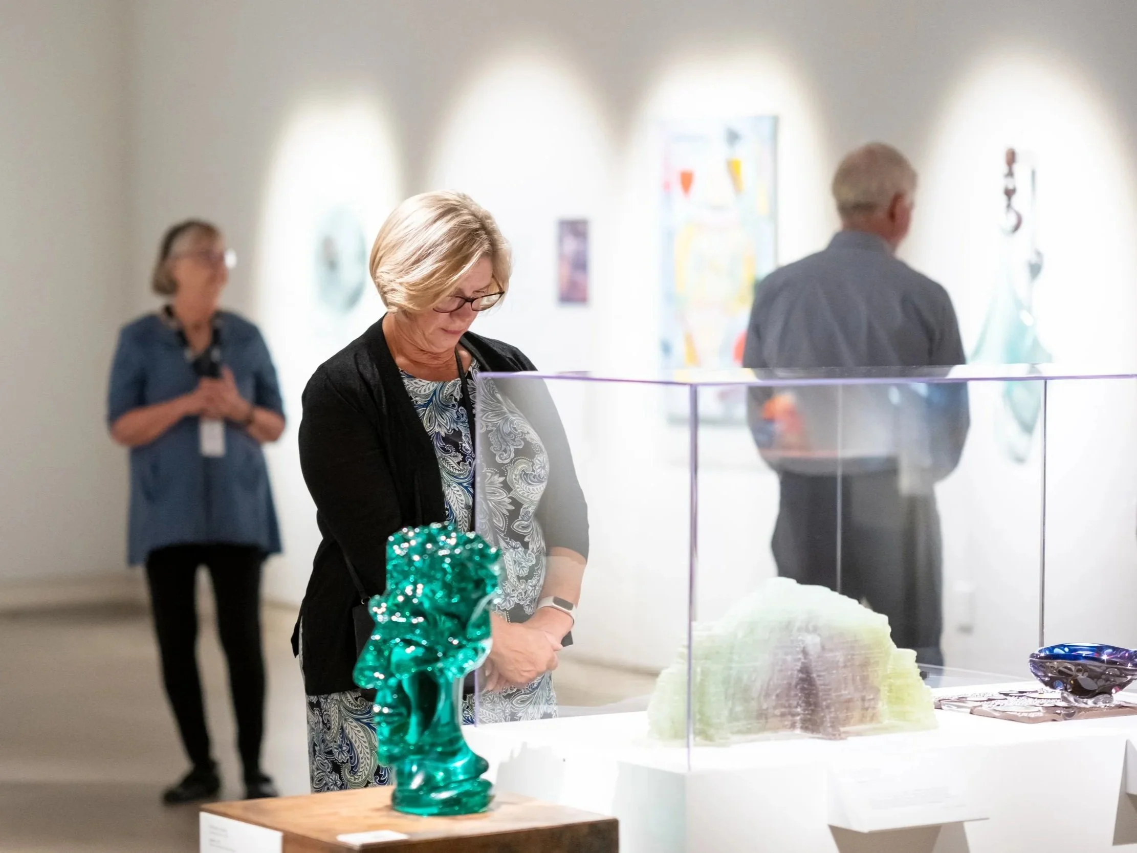 A woman observing glass sculptures with artwork on the wall behind her at an art exhibition.