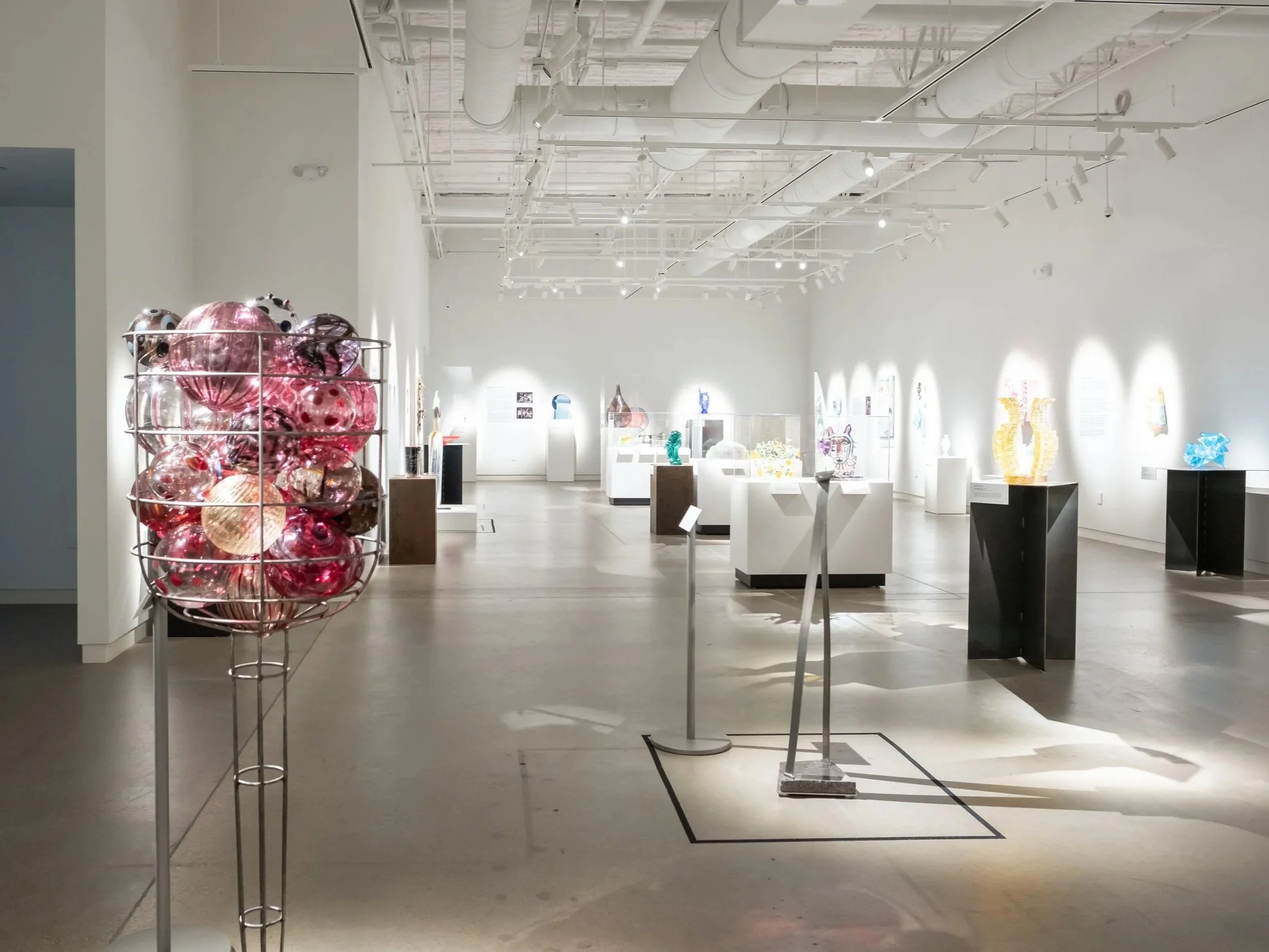 Interior of an art gallery or museum with various glass sculptures and artworks on display, illuminated by track lighting ceiling fixtures.
