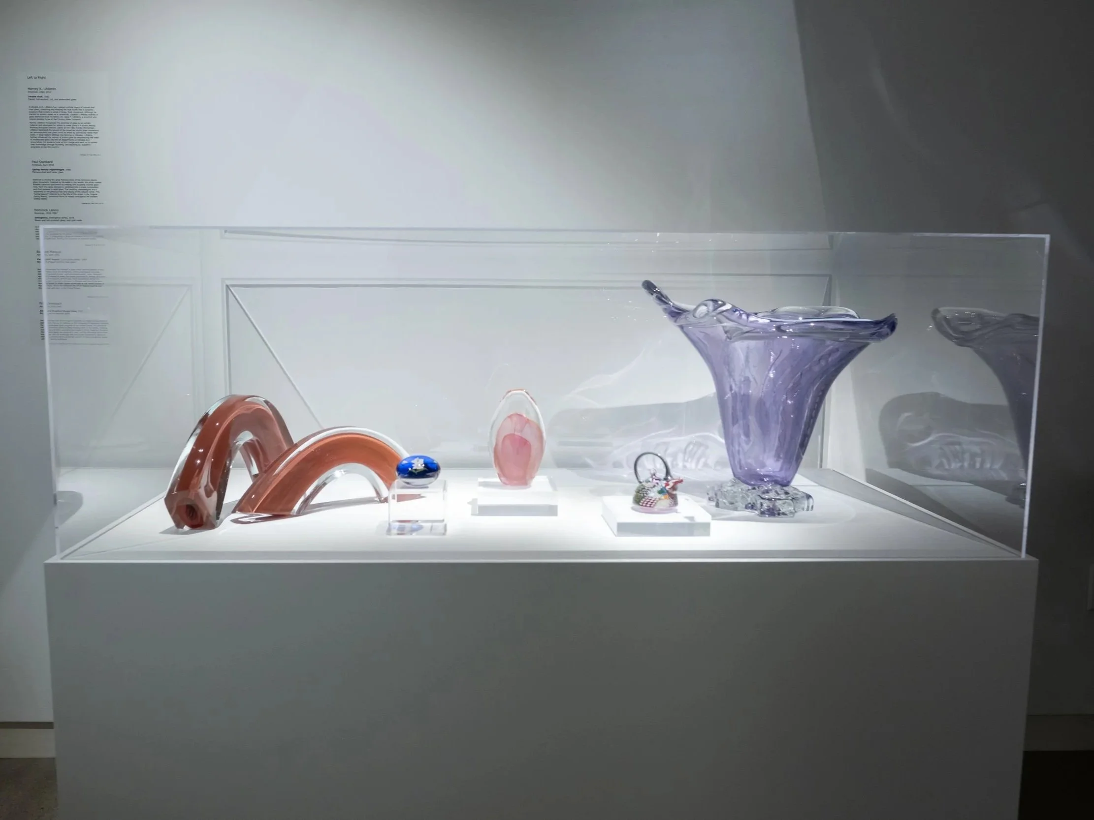 Display case with various glass art sculptures, including a purple vase, pink and red glass arcs, and small decorative objects, in a museum or gallery setting.