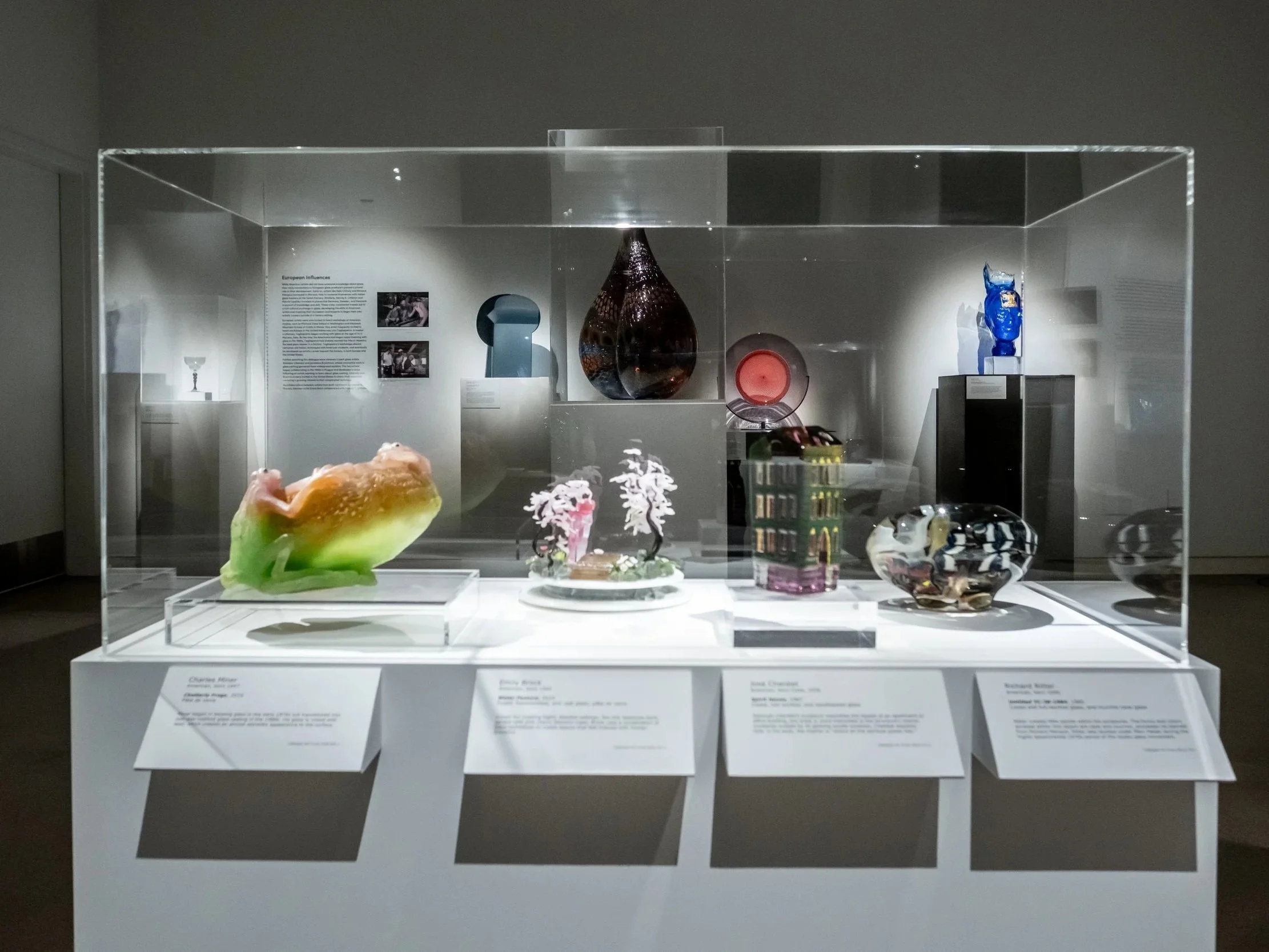 Museum display case with various glass and ceramic art objects, including a multicolored piece in the foreground and several smaller items and informational plaques behind it.