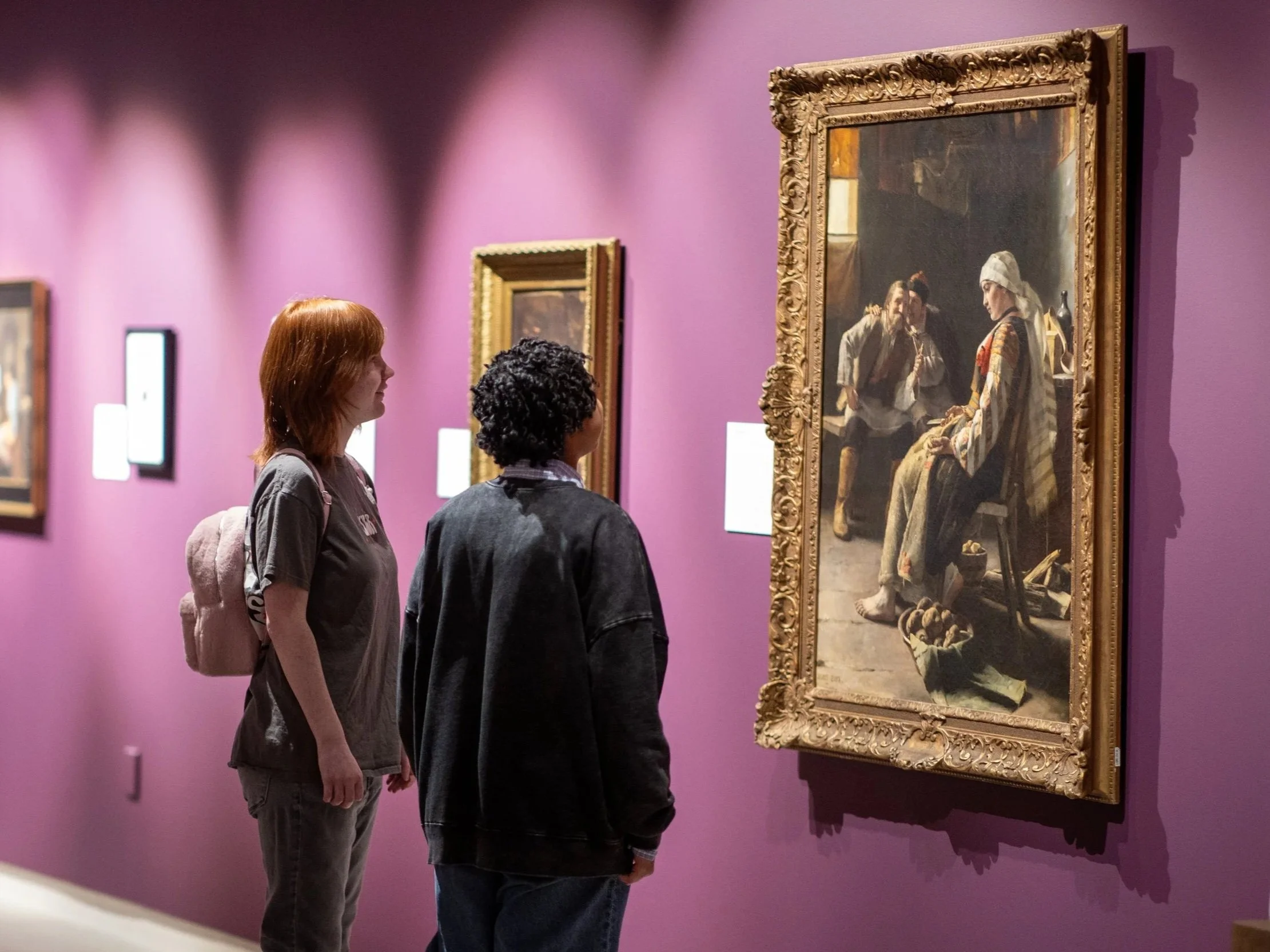 Three people standing in front of a large framed painting in a museum with purple walls, looking intently at the artwork.
