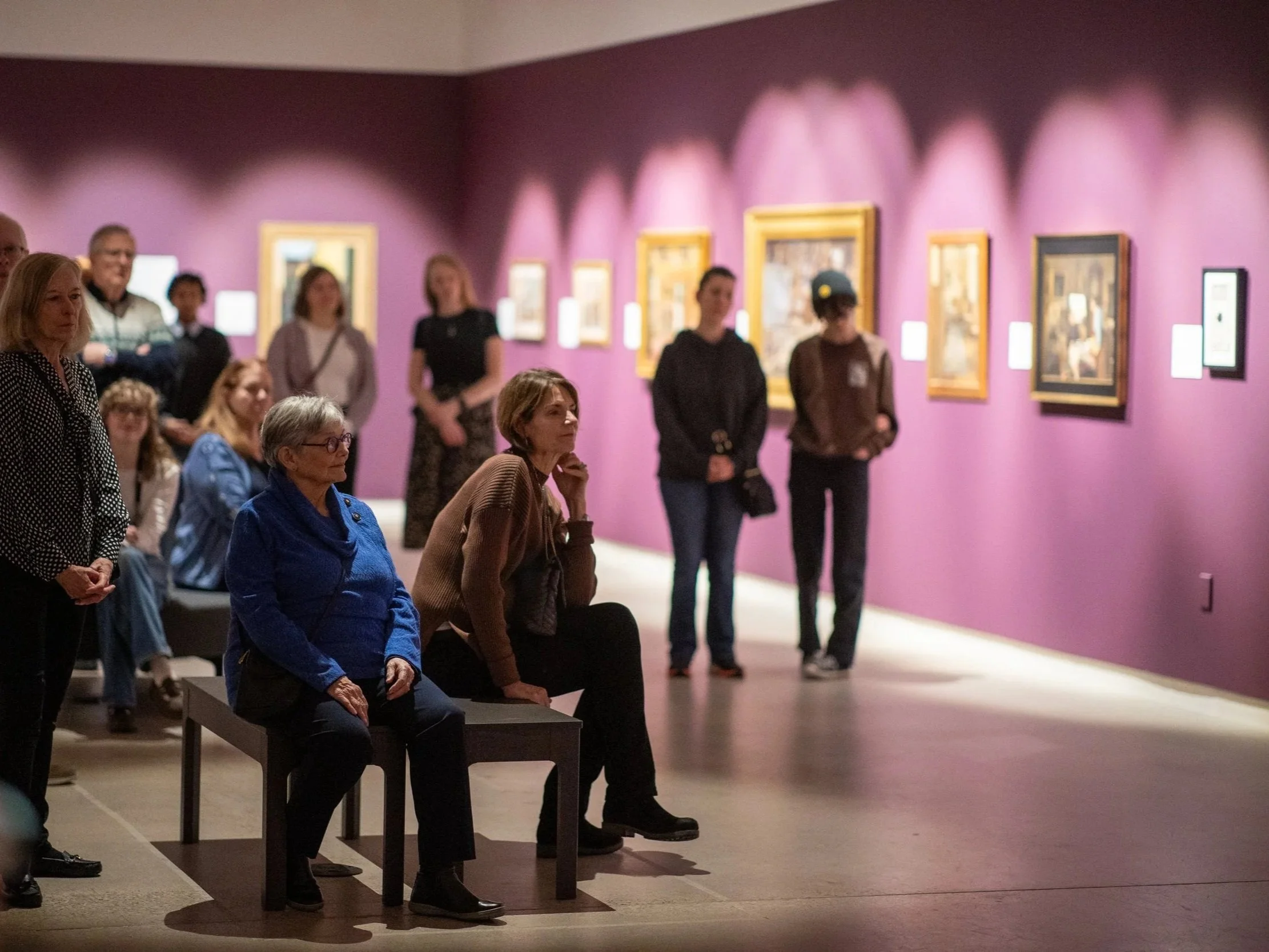 A group of people in an art gallery observing paintings on a purple wall. Some are sitting, some standing, and others are looking at the artwork.