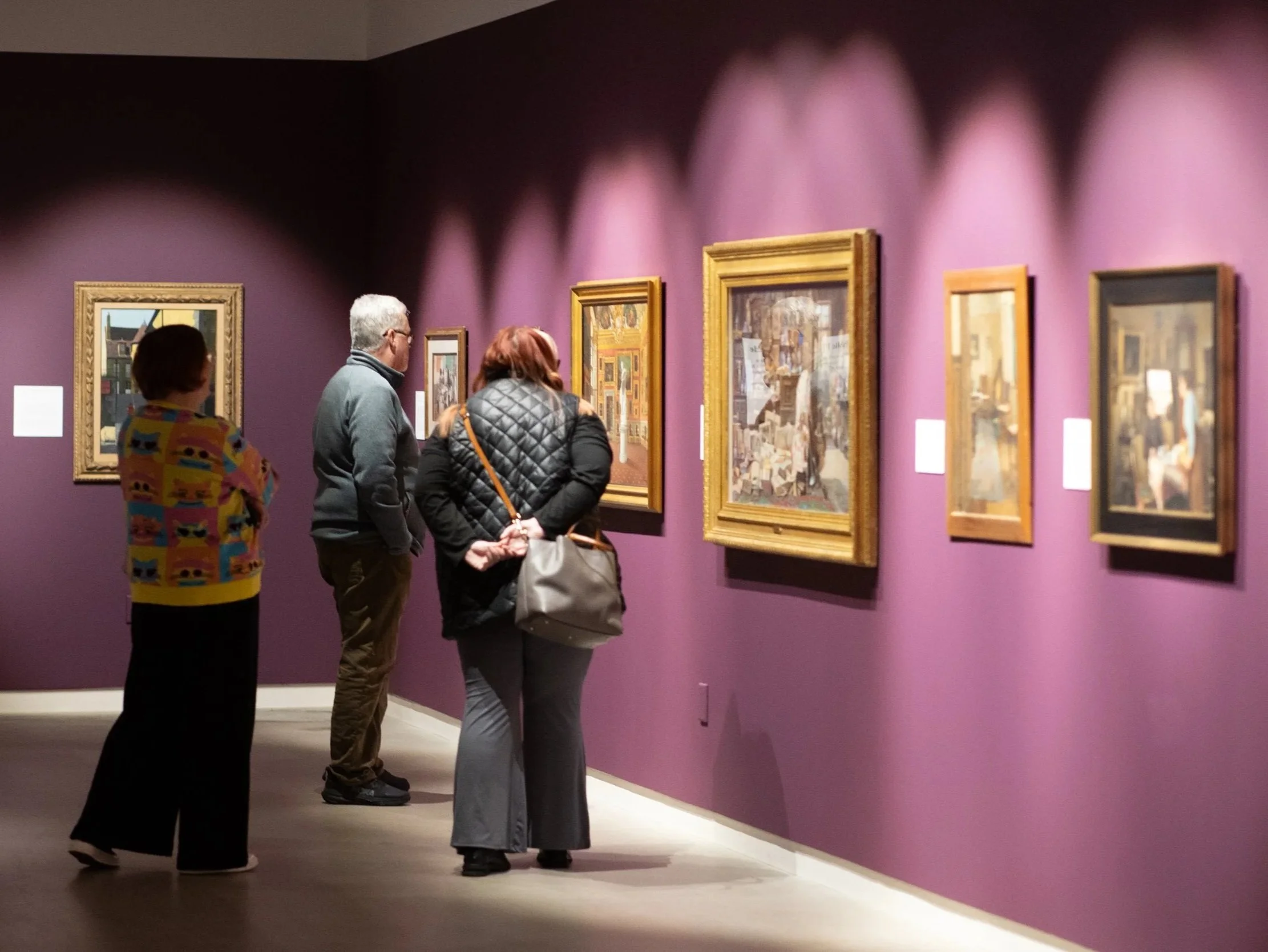 Three people are viewing framed artwork on a purple wall in an art gallery, with two women and one man standing close together, engaged in viewing the paintings.