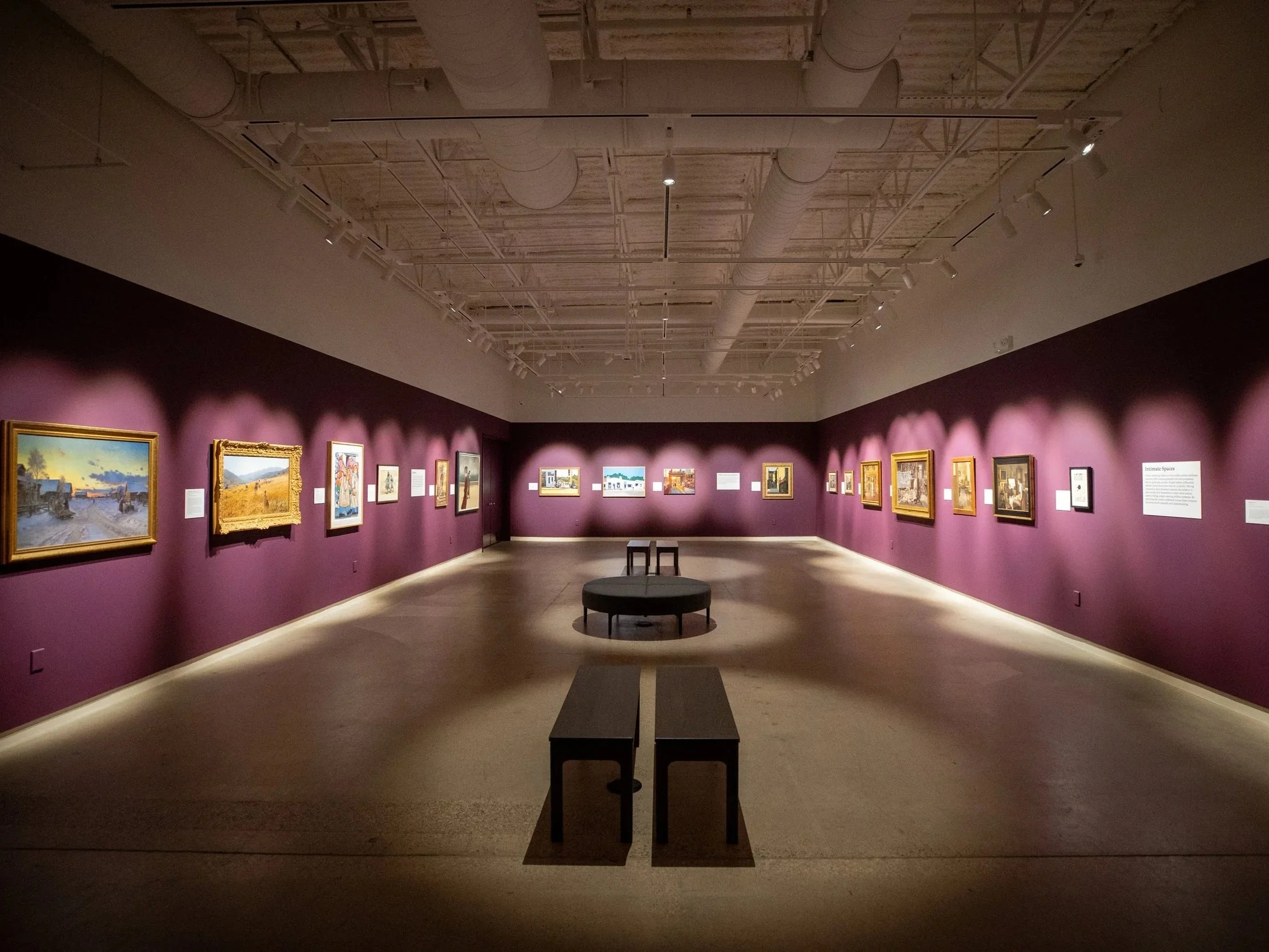 An art gallery with paintings displayed on purple walls, track lighting on the ceiling, and benches in the center of the room.