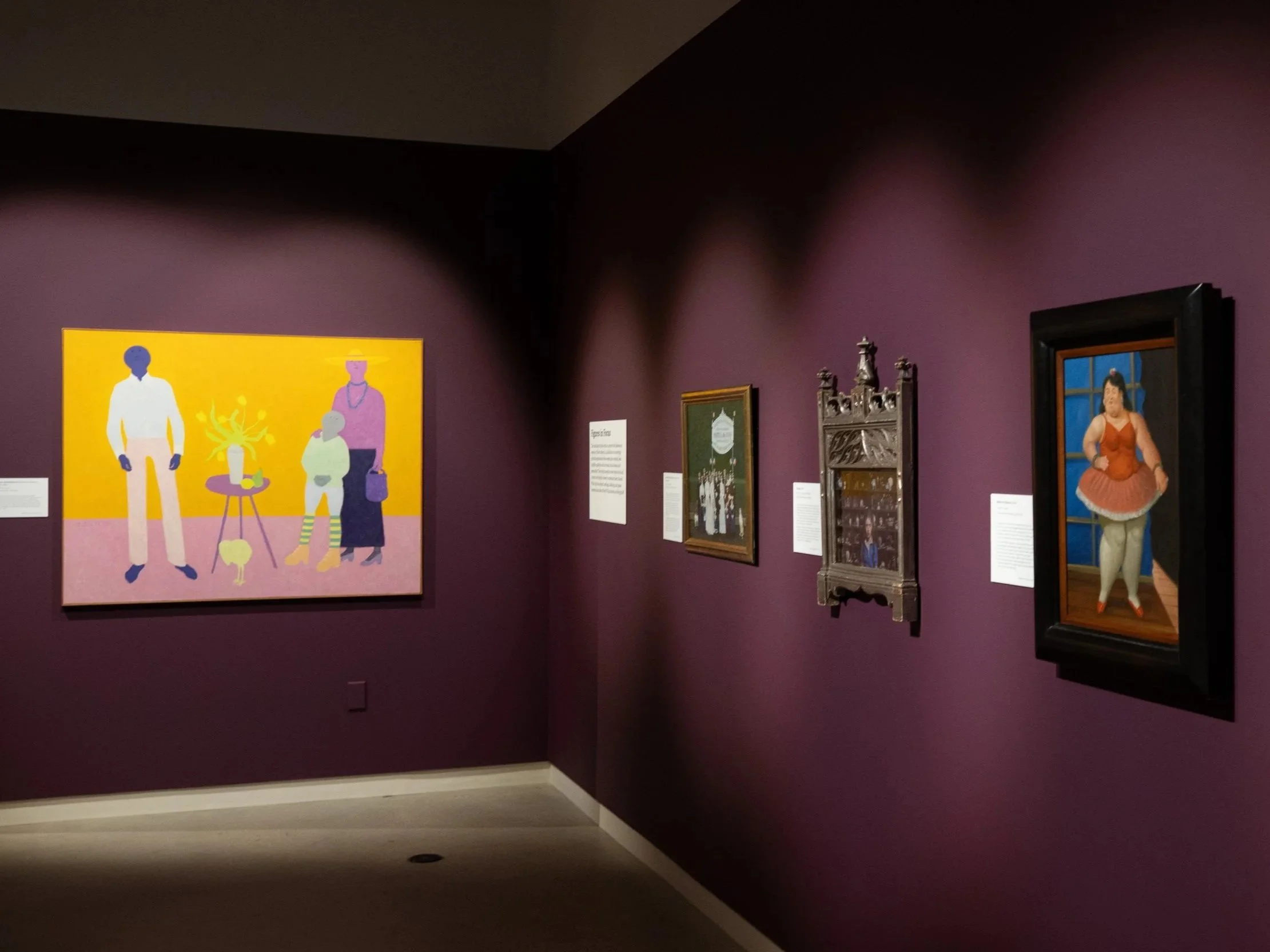 Museum gallery with purple walls displaying framed artworks including a colorful illustration of people and other paintings; part of a sculpture is visible.
