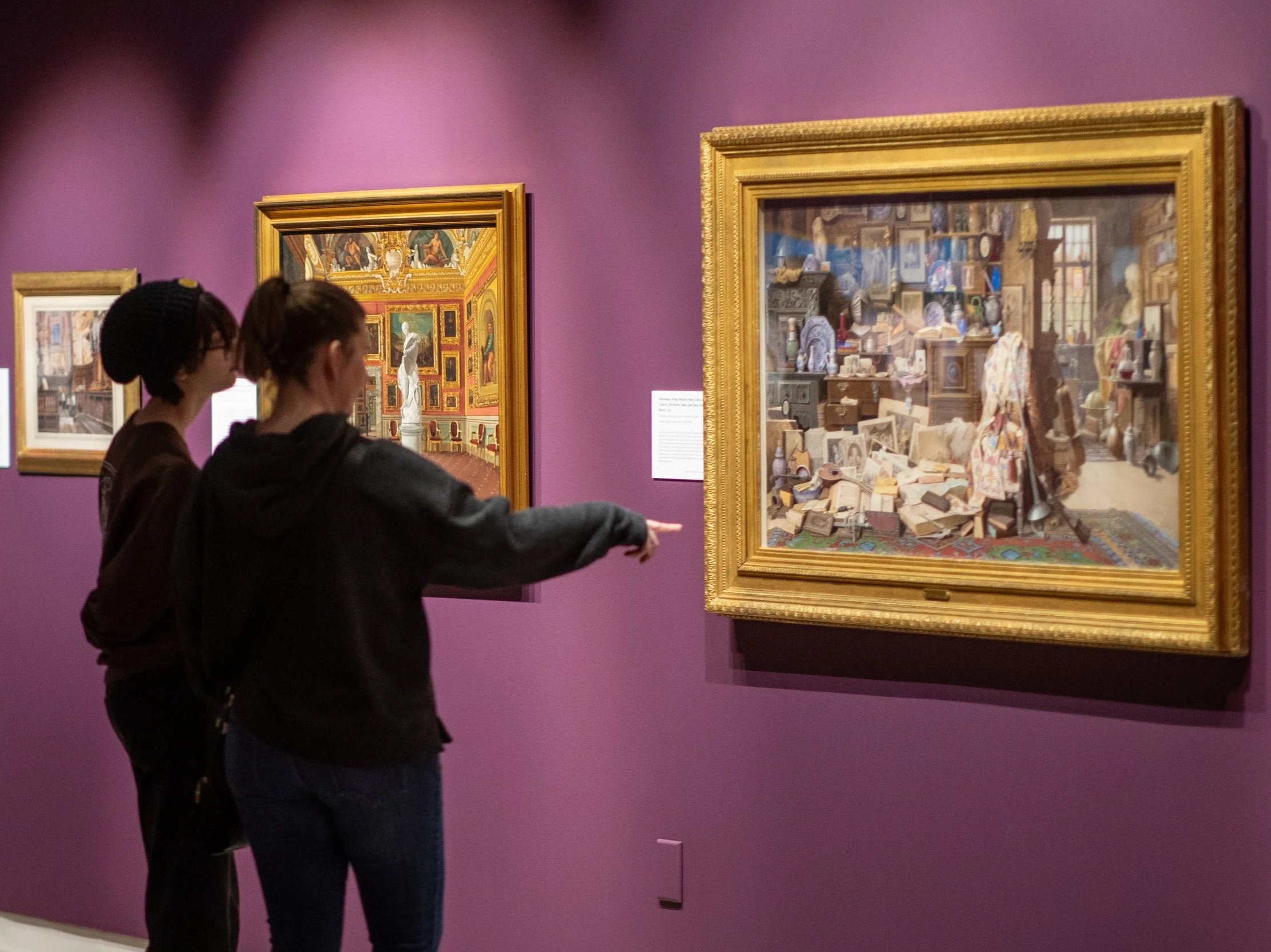 Two women are examining framed artwork in a gallery with purple walls. One woman is pointing at the artwork, which contains an interior scene filled with various objects and a cluttered room.