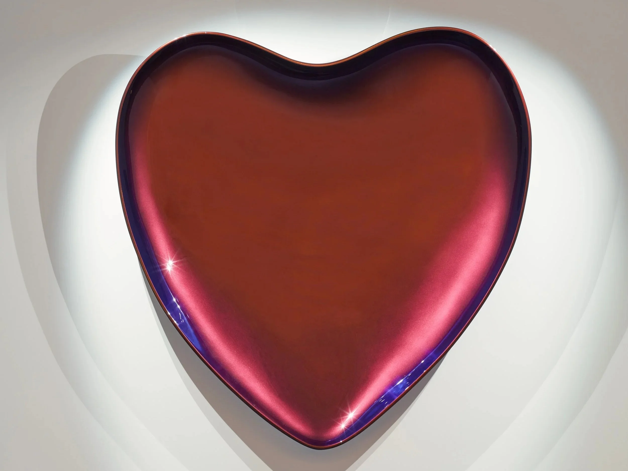 A shiny, metallic, heart-shaped object with a dark red color, reflecting light on its surface, against a white background with soft shadows.