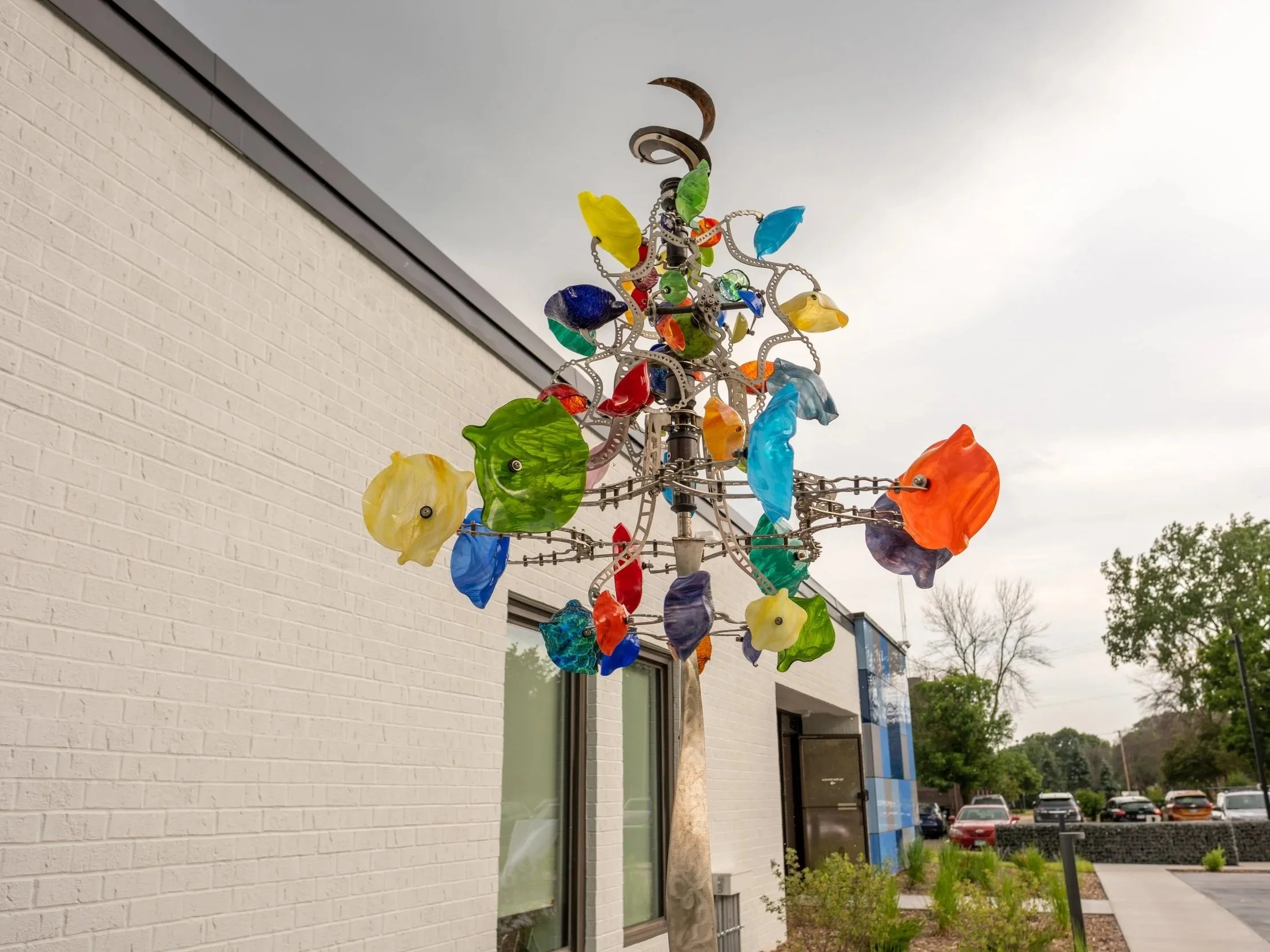 Colorful glass wind sculpture mounted outdoors near a modern building with white brick wall and large windows, adjacent to a parking lot with cars, trees, and cloudy sky.