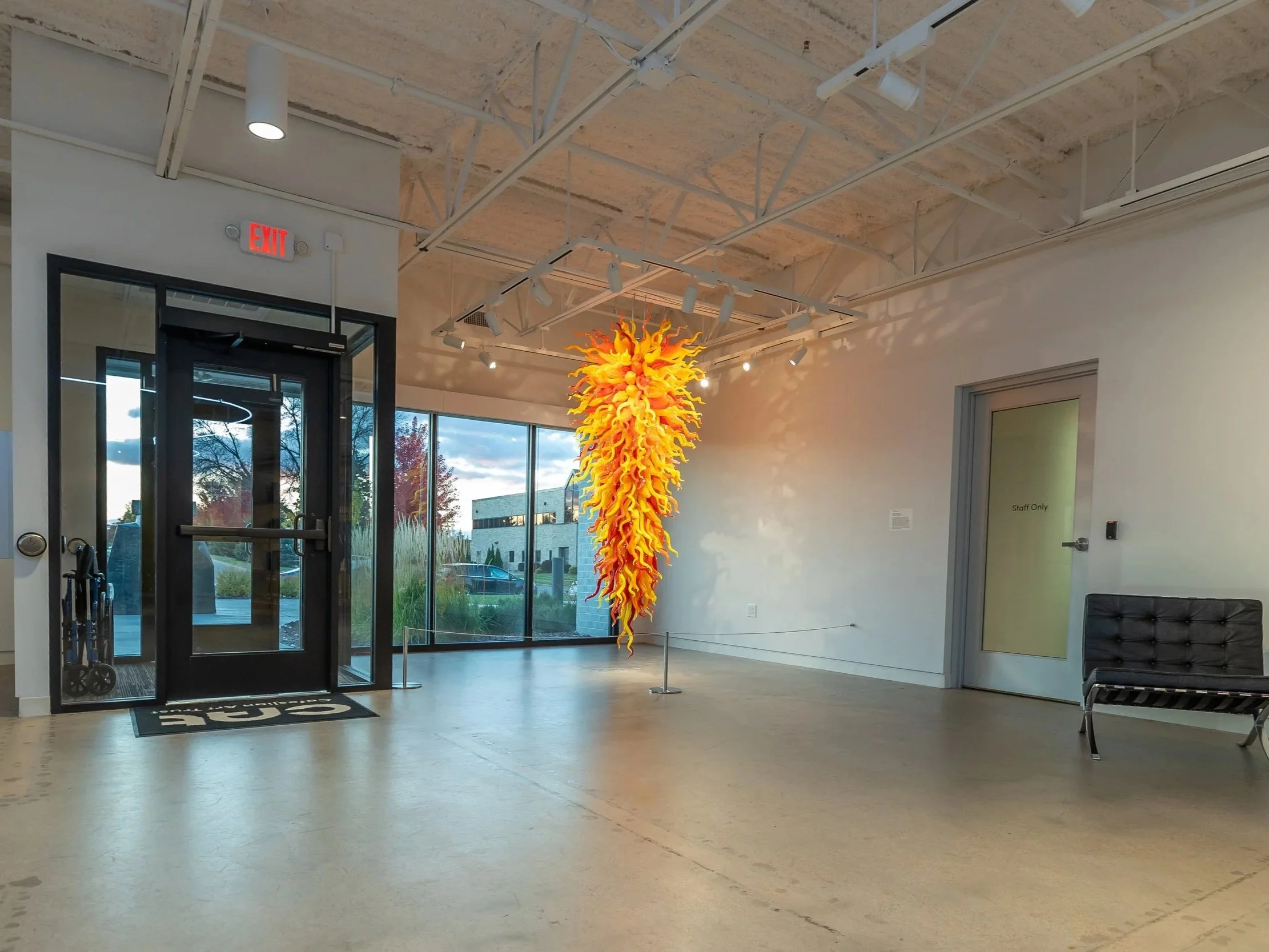 Interior of a building with a large hanging glass sculpture in bright yellow and orange colors. The space has large windows, a glass door, a black bench, and an 'Exit' sign above the door.