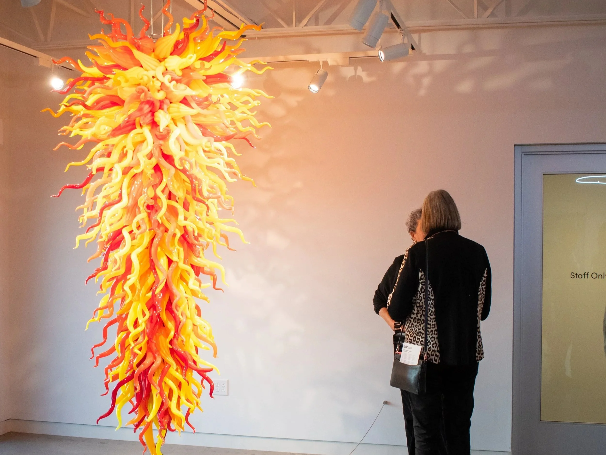 A colorful glass sculpture resembling a fiery sunburst with yellow, orange, and red elongated tendrils hanging from a ceiling in an art gallery. Two people are observing the artwork, one with curly hair and the other with straight hair, wearing dark 