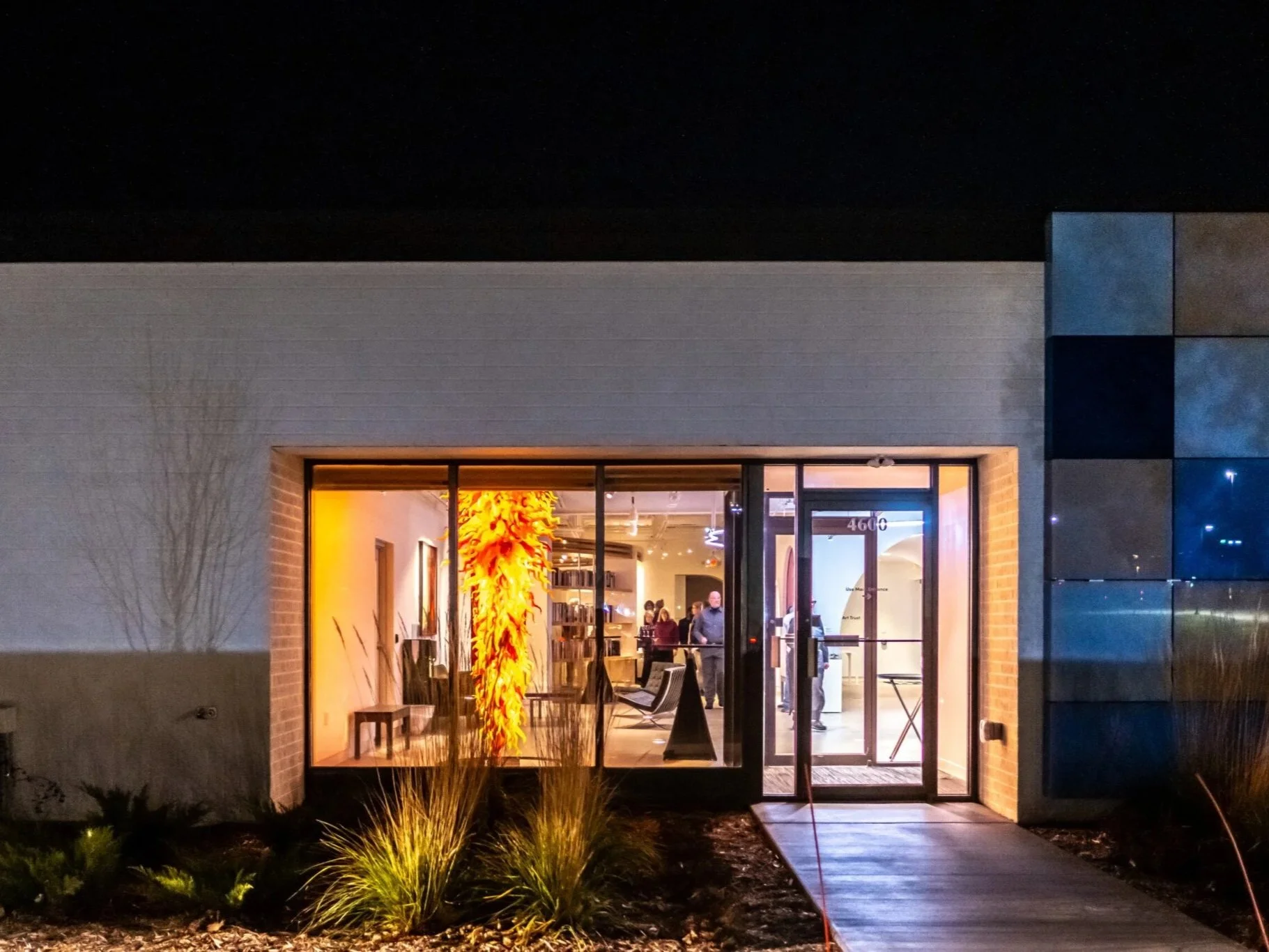 Night view of a modern building with large glass windows and an illuminated interior, featuring an abstract orange and yellow sculpture inside, with some outdoor landscaping in the foreground.