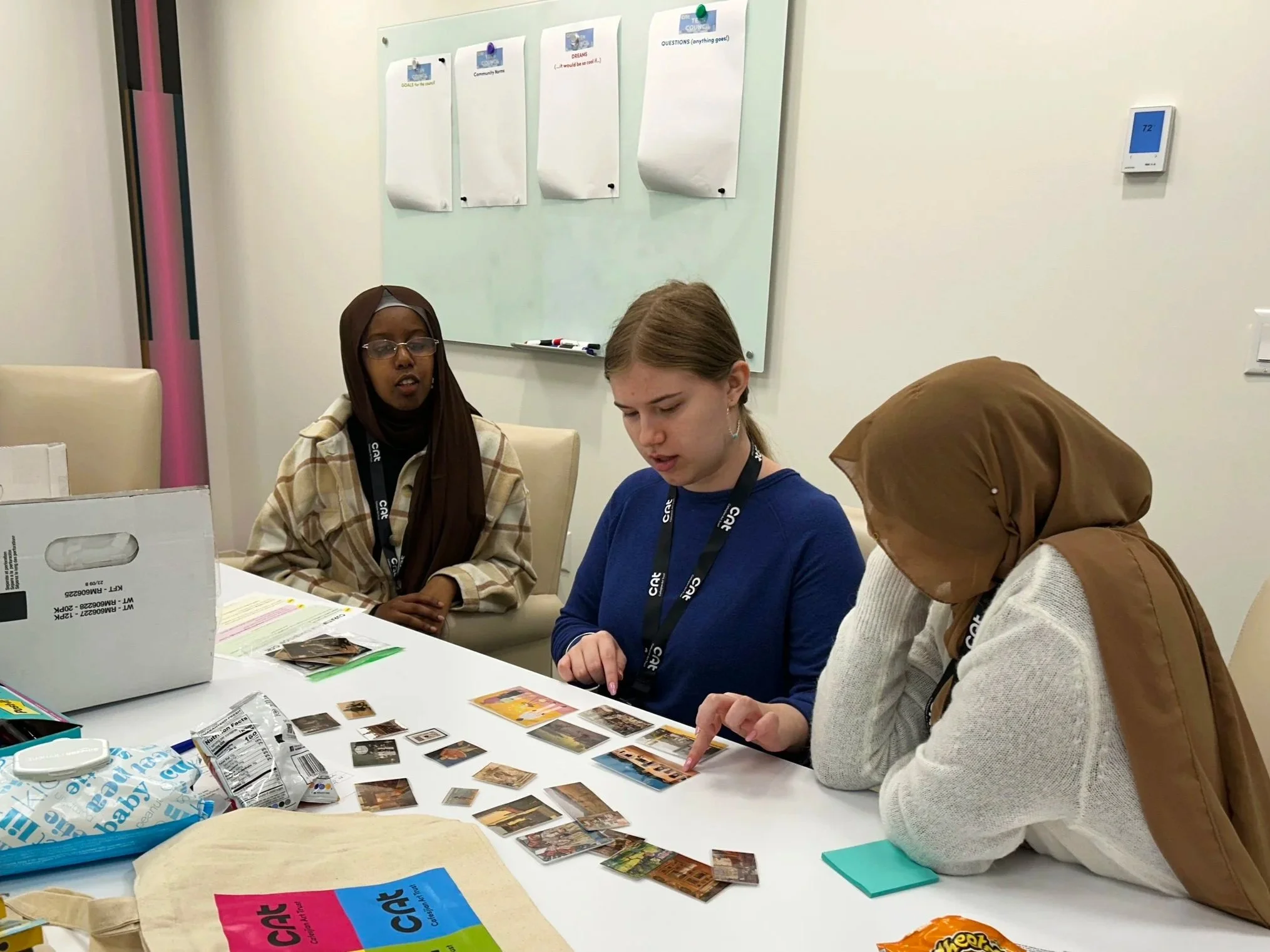 Three teenagers working together in a conference room to arrange miniature paintings on the table.