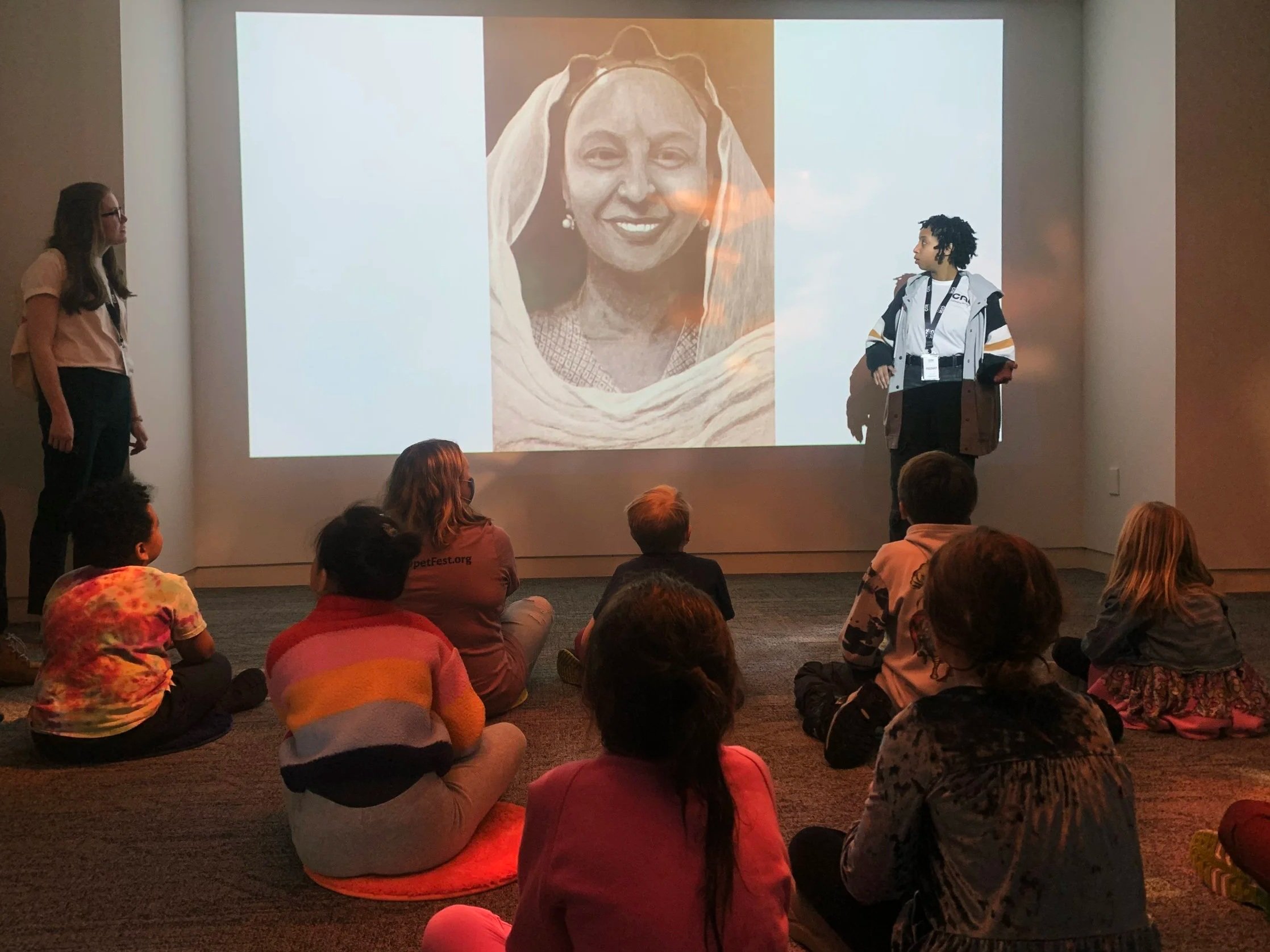 A teenager stands in front of a group of children seated on the floor, they are looking at a drawing of a woman projected on the wall.