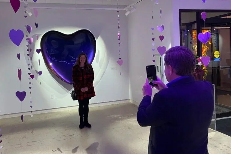 A person poses in front of the CAT's Purple Heart sculpture by Tommy Tanggara while someone takes their photo. Purple heart streamers are hung from the ceiling. 
