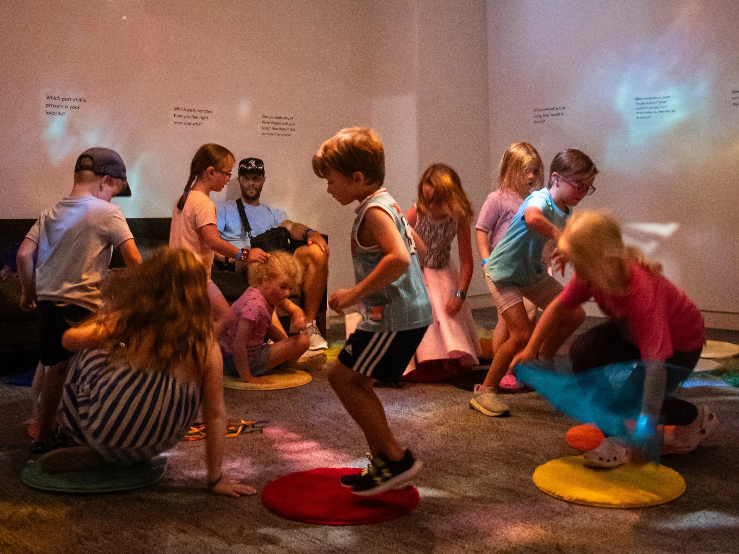 A group of children move in a circle, stepping onto colorful carpet circles. 