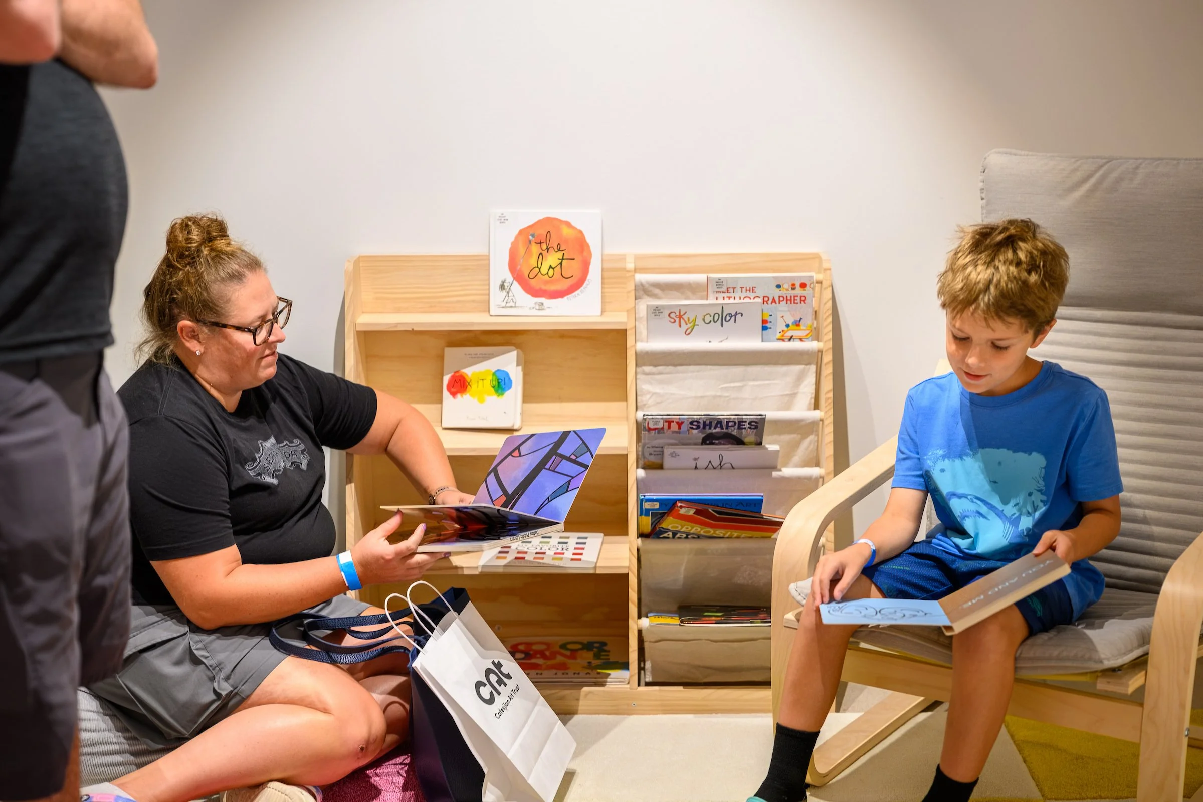A woman sitting and reading a colorful book to a young boy, who is sitting in a wooden chair, in a children's library or reading area with bookshelves and children's books visible in the background.