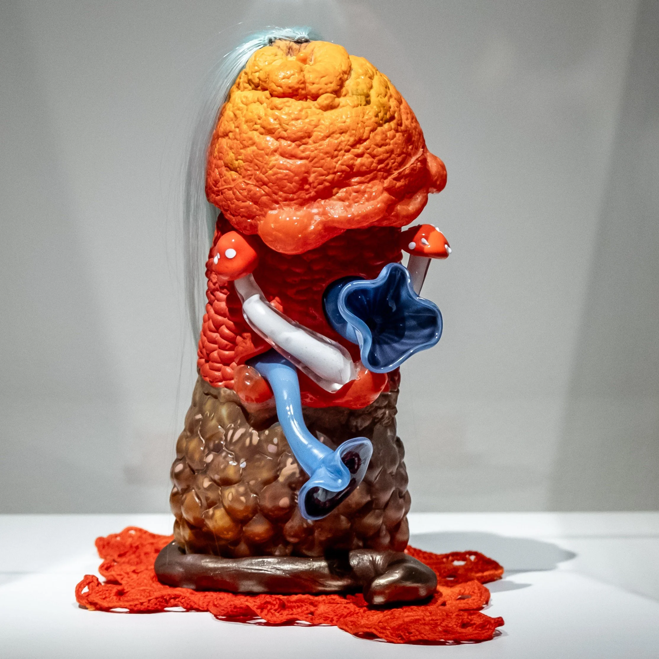 Abstract sculpture with textured orange, blue and brown surface, protruding mushrooms, and white hair.