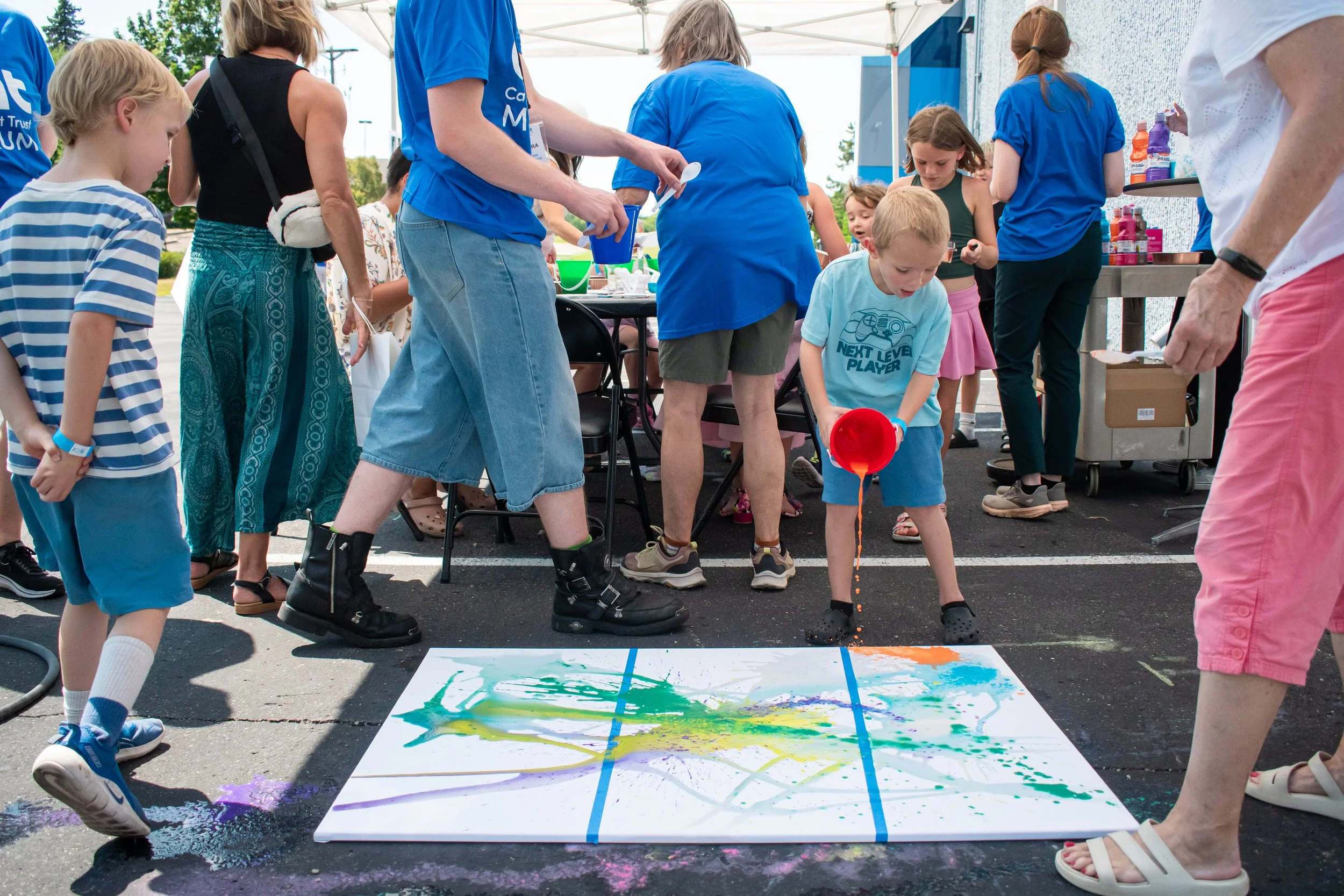A group stands around a canvas on the ground while a child pours a bucket of paint onto the canvas. 