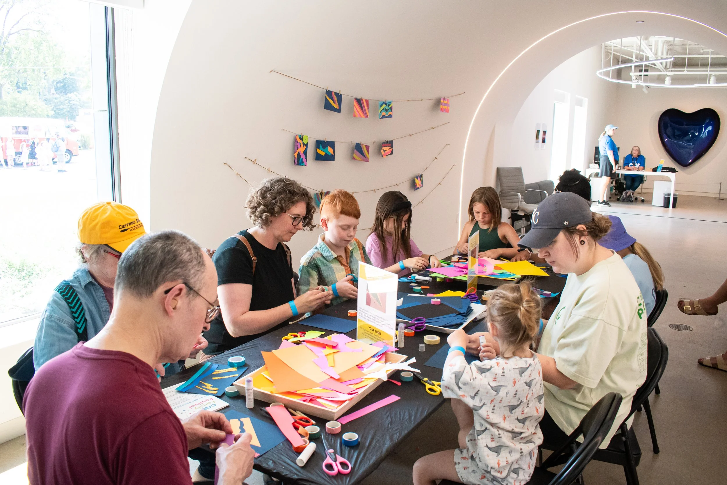 A group of people of varying ages sit around a table and make art with colorful paper. 