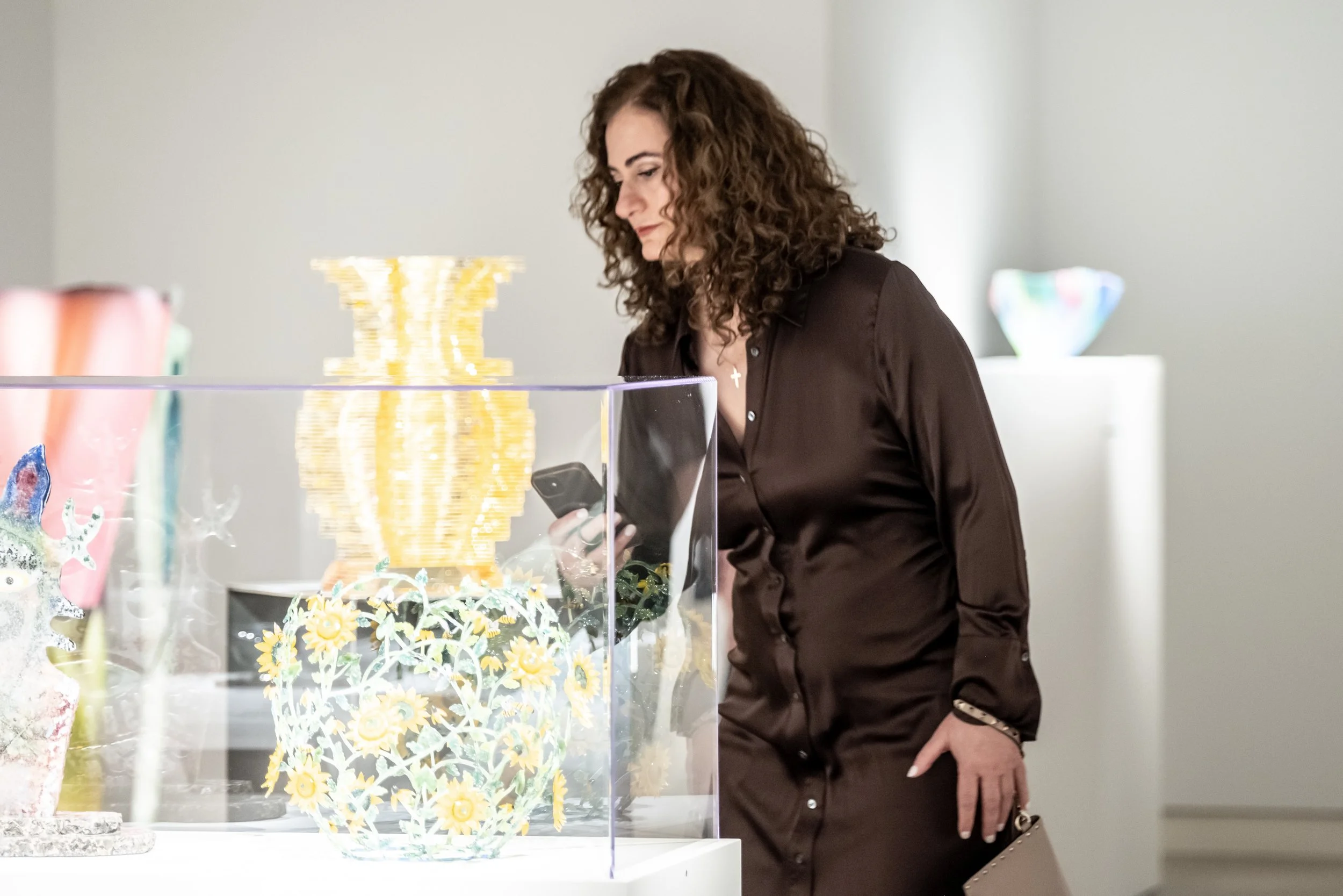 A woman with curly brown hair wearing a dark blouse looks at a glass display case with intricate yellow floral artwork in an art gallery.