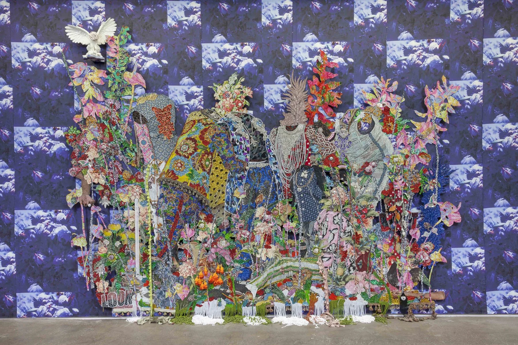 A mixed media art installation featuring embroidered and fabric artwork of elephants, set against a deep blue floral backdrop in a gallery space.