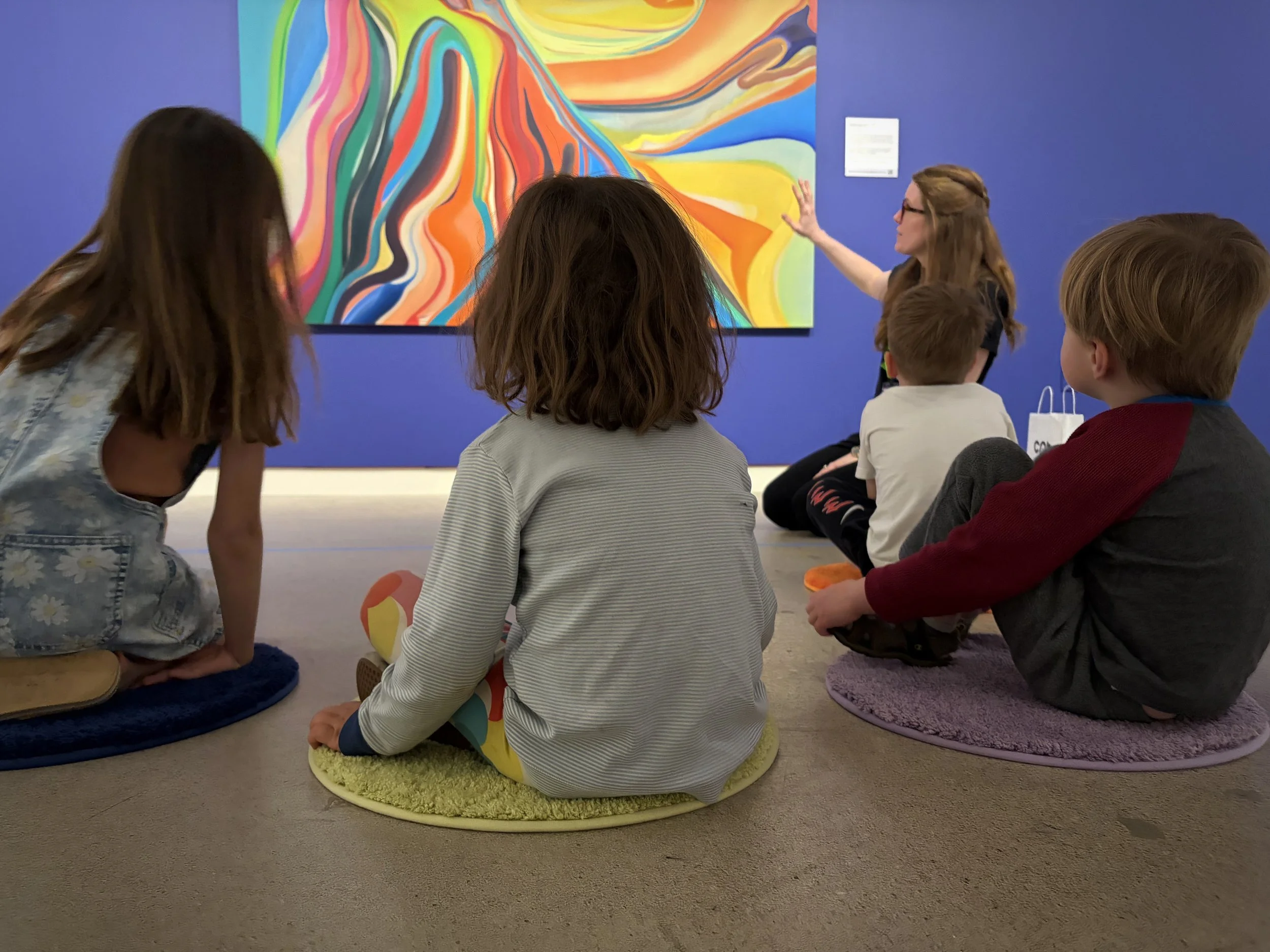 Children seated in front of a colorful abstract painting listening to an educator.