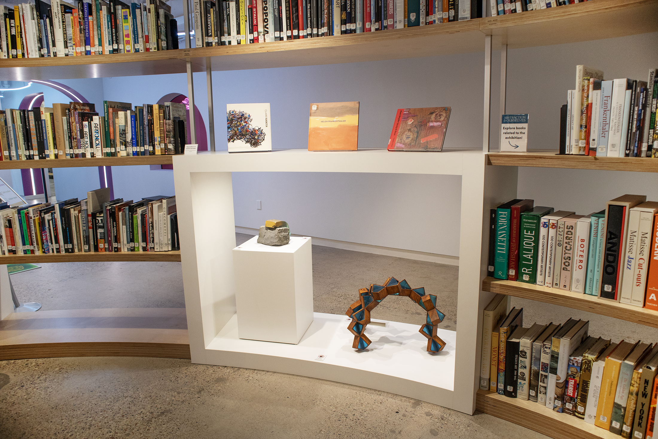 A library or bookstore with books on wooden shelves. There is a white display table in the center with three small books and two geometric wooden sculptures.