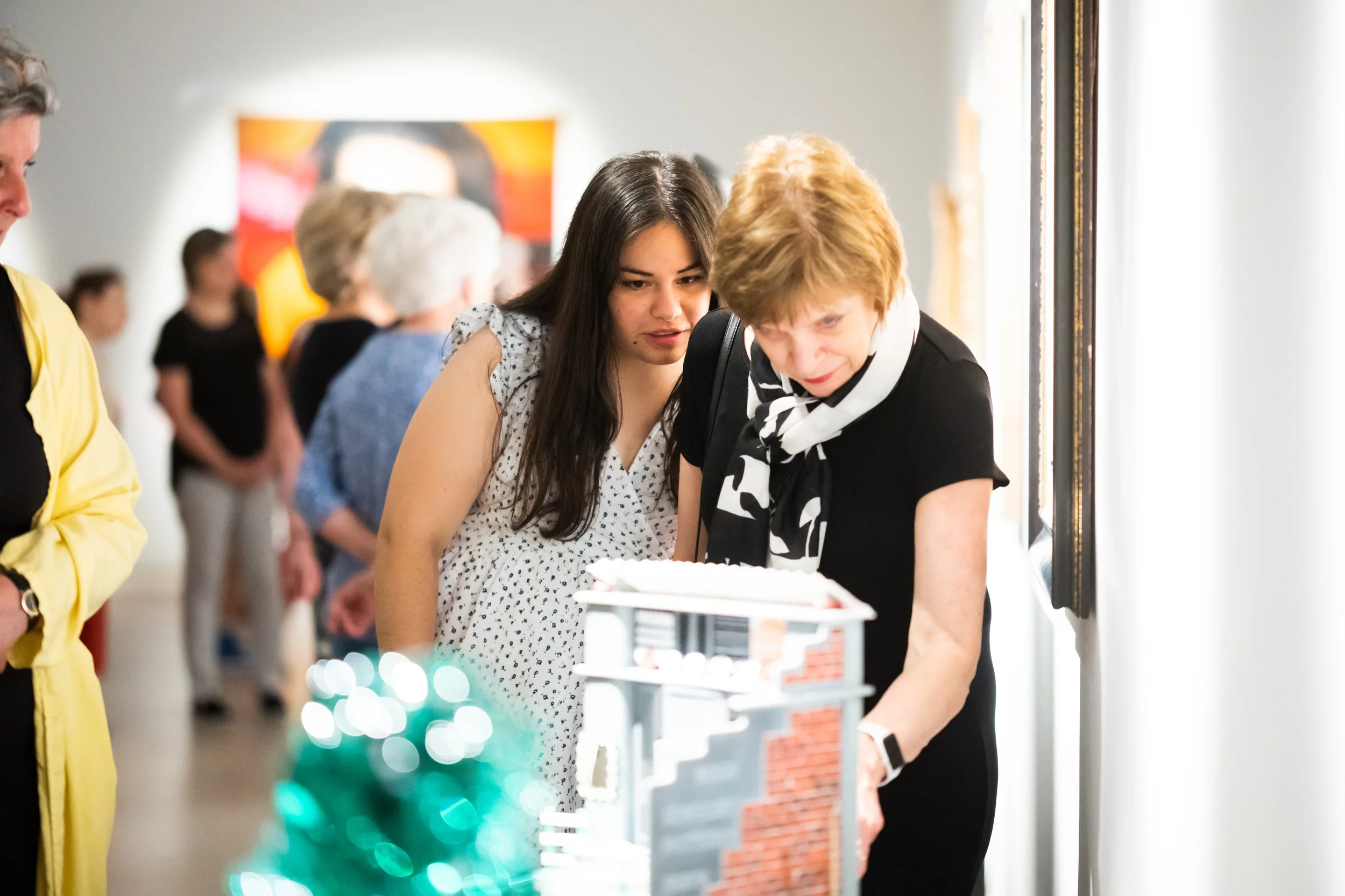 Two women closely examining a display or artwork at an indoor exhibition or gallery while other people are in the background.