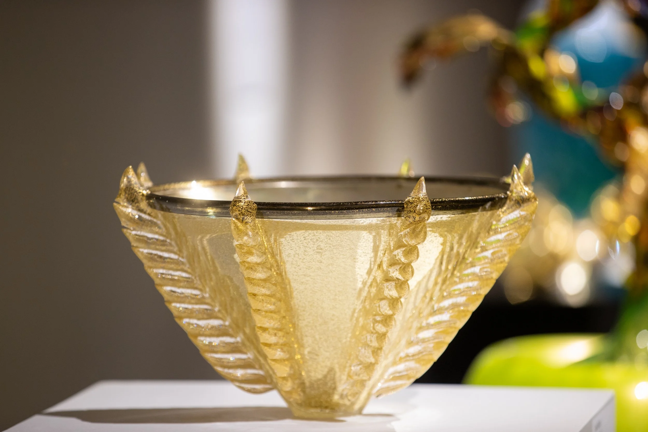A decorative golden glass bowl with a textured pattern, sitting on a white surface with blurred colorful ornaments in the background.