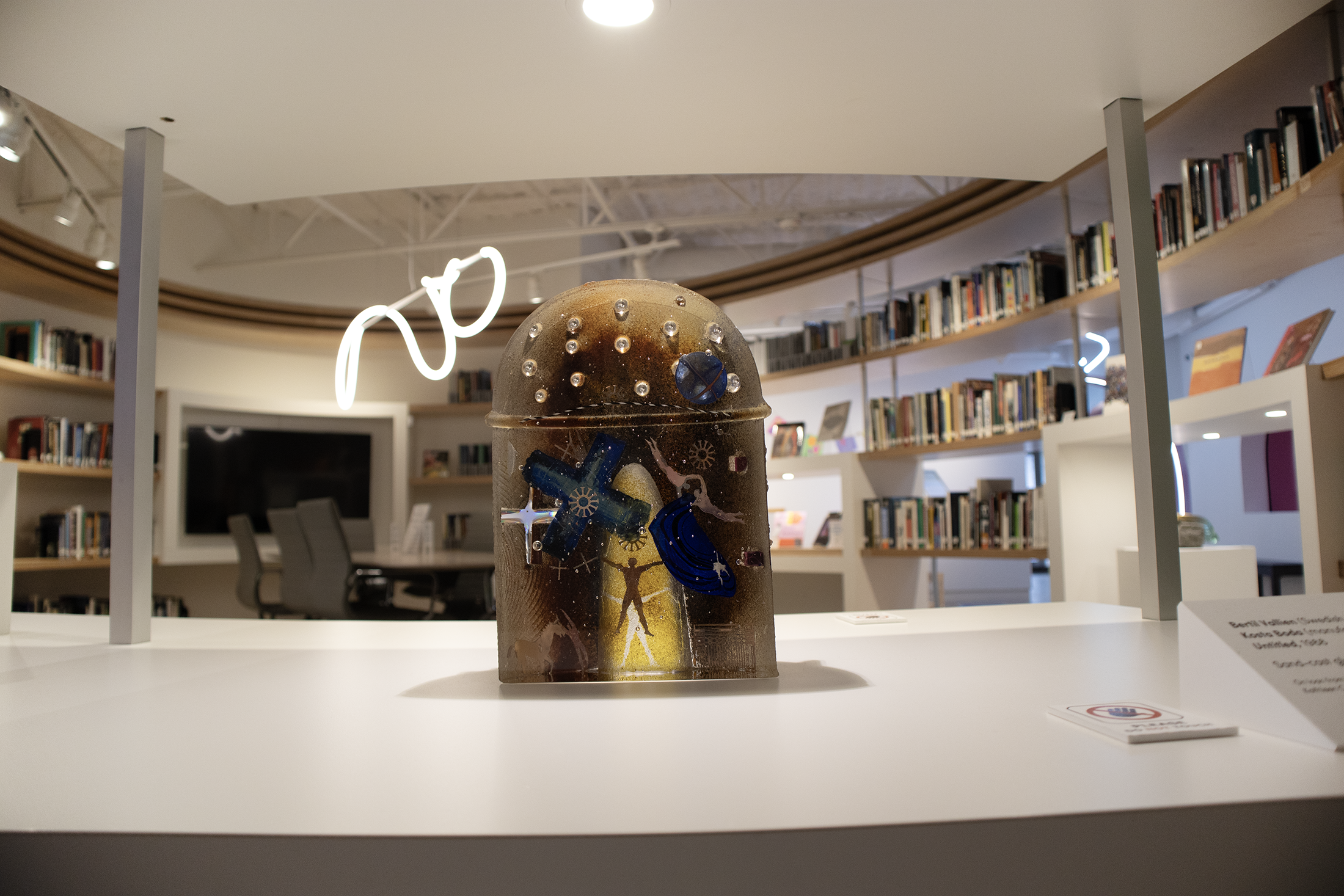A decorated, semi-transparent, cylindrical object with images and symbols, placed on a white table in front of bookshelves in a room with modern decor.