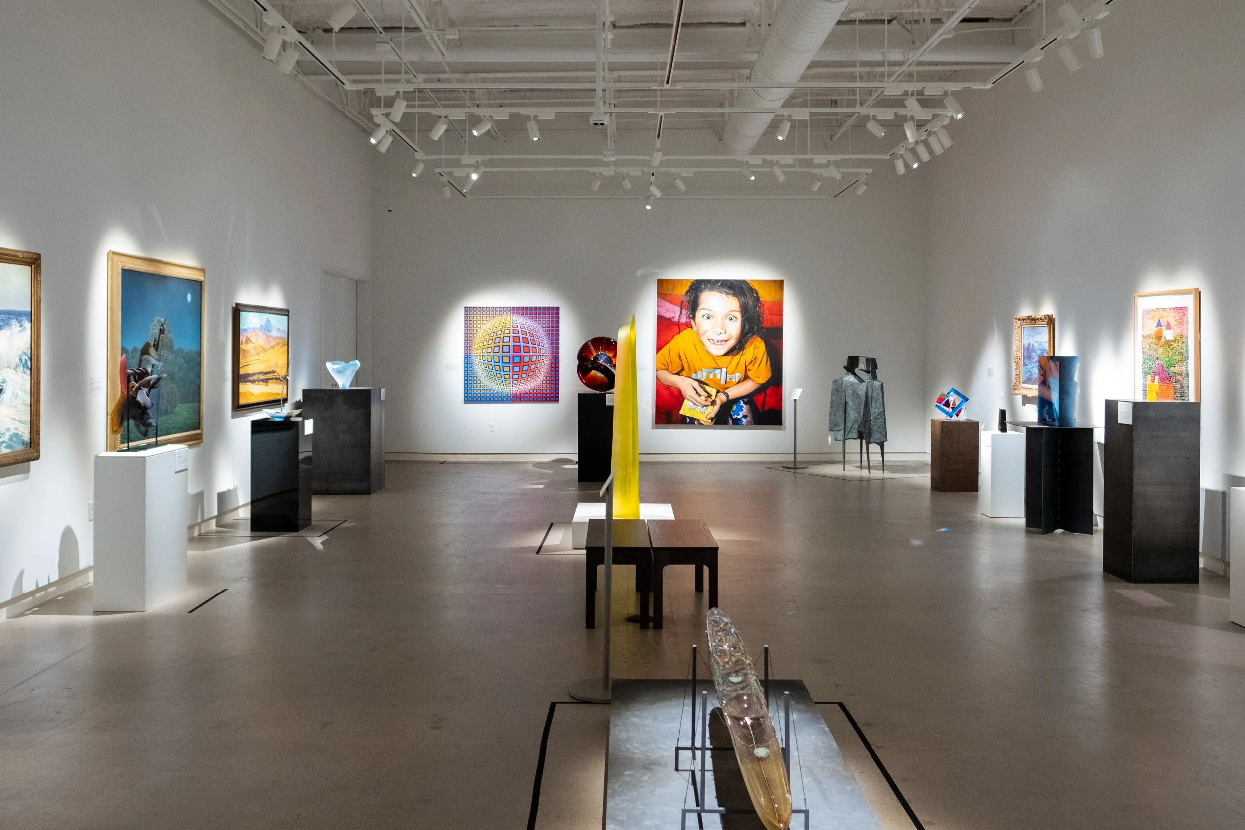 An art gallery featuring various colorful paintings and sculptures displayed on pedestals and walls, with track lighting illuminating the artwork.
