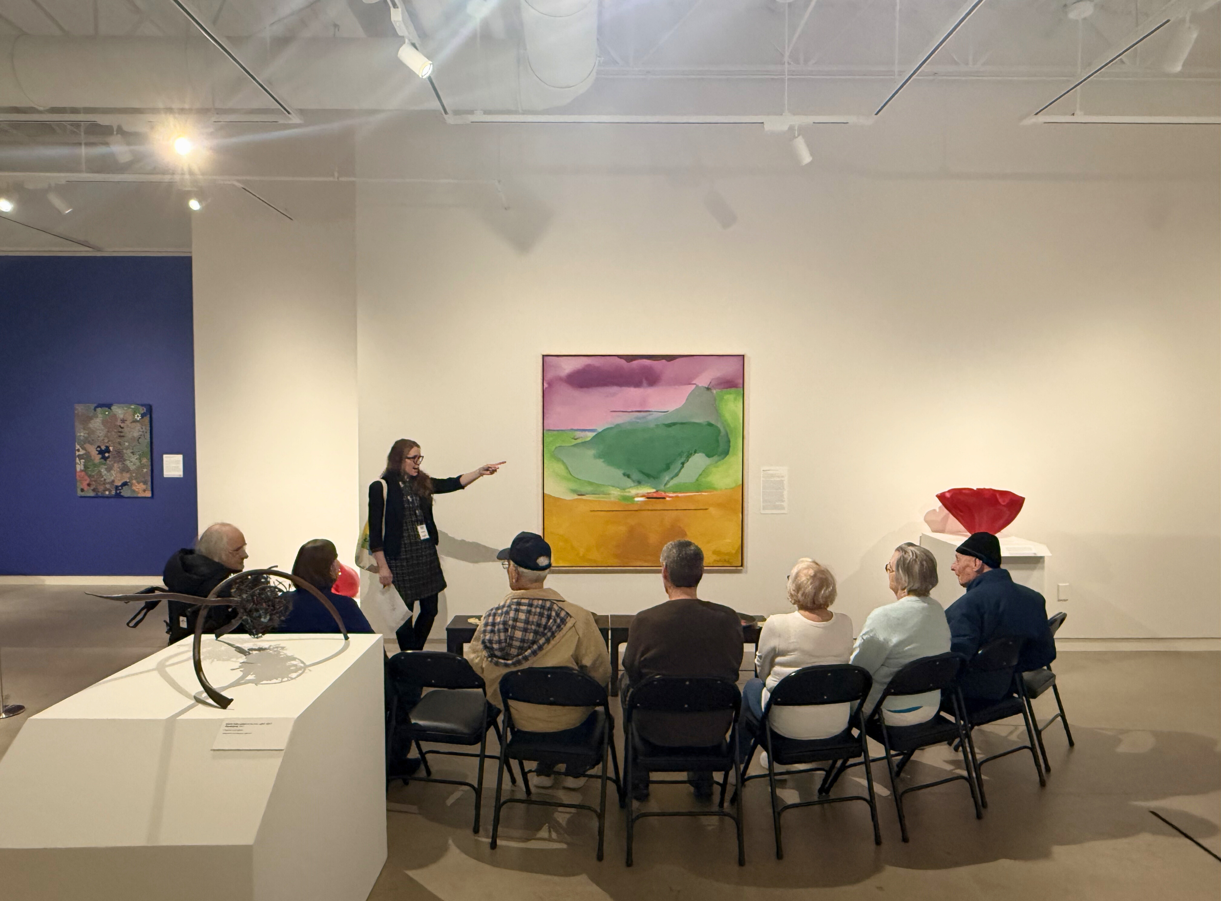 An female educator pointing at a painting, leading a discussion for a group of seated elderly participants.