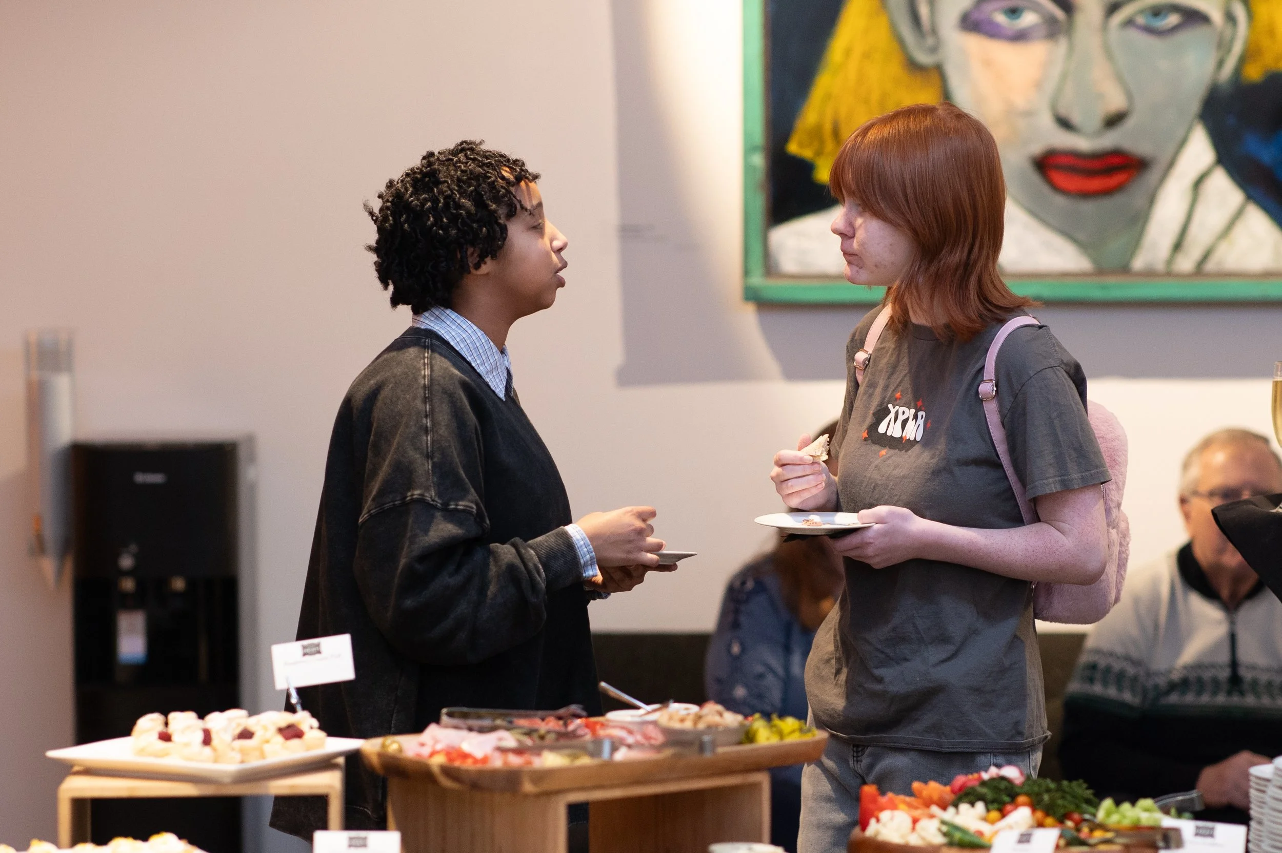 Two teens talk and enjoy food, standing by a table with an array of appetizers. 