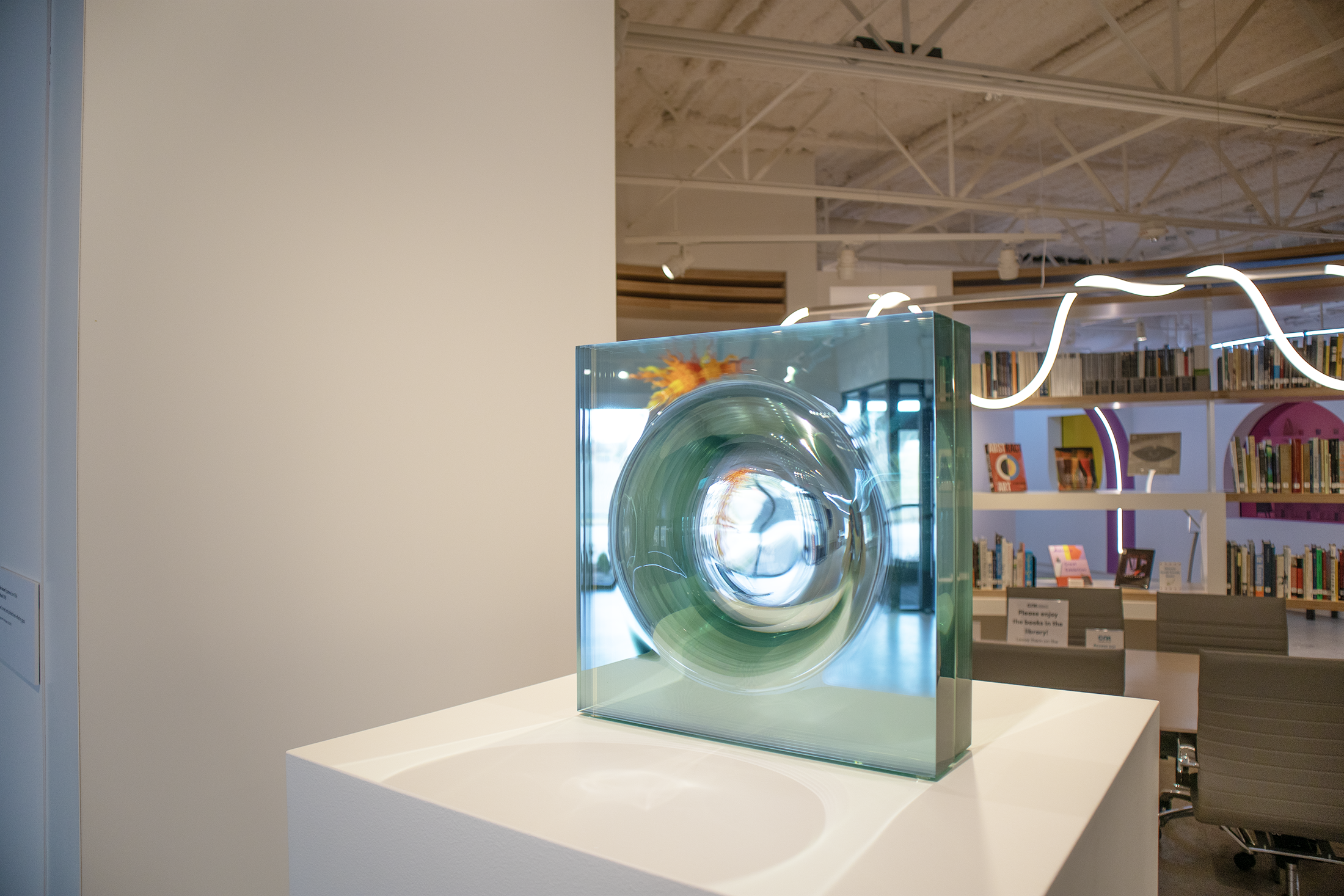 A glass art sculpture with a swirling pattern, displayed on a white pedestal in a modern library or gallery space.