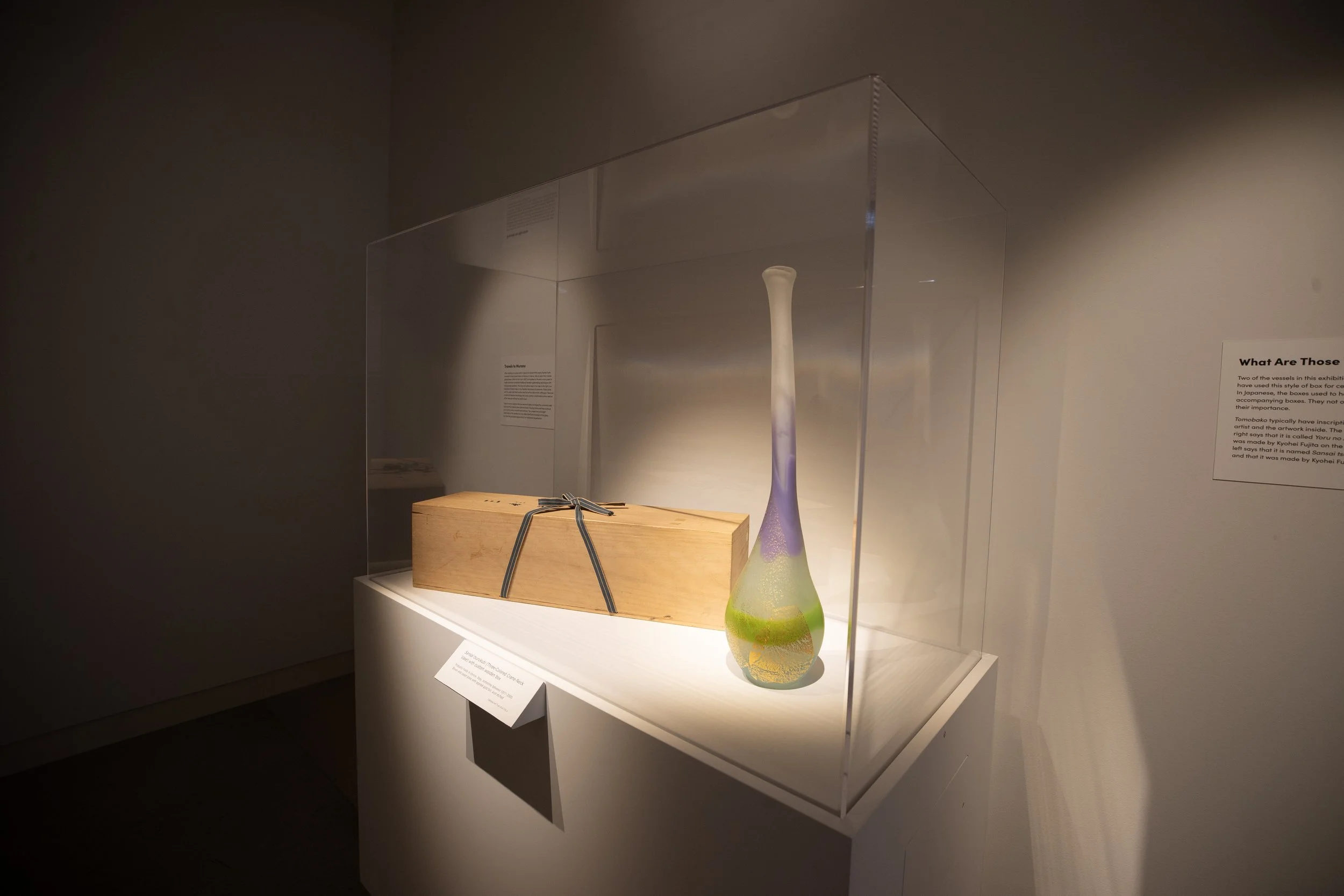 A display case containing a tall, colorful glass vase with a gradient of colors from purple at the top to green at the bottom, and a wooden box tied with a gray ribbon.