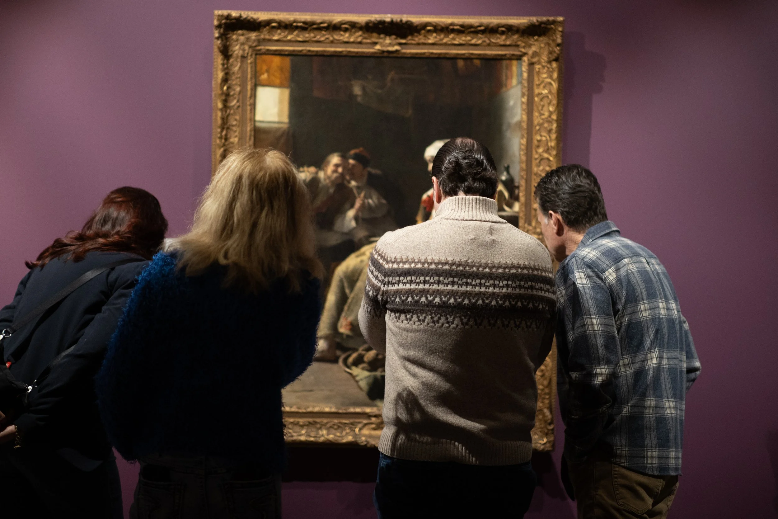Adults gather to look at a painting in an ornate frame.