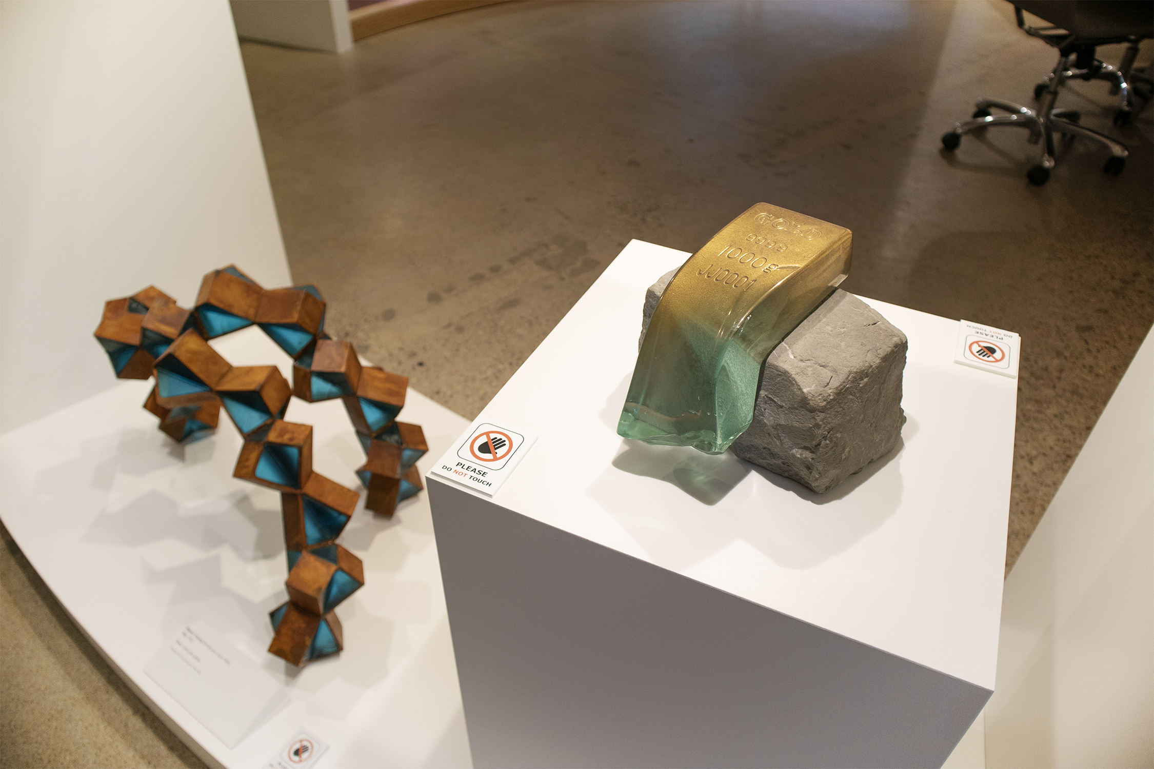 Exhibit display with a geometric, open-dodecahedron sculpture with blue interior on the left, a rectangular rock with a mounted greenish metallic weight marked "1000 lb" on a bronze-colored base in the center, and a no-touch sign on the right, in a g