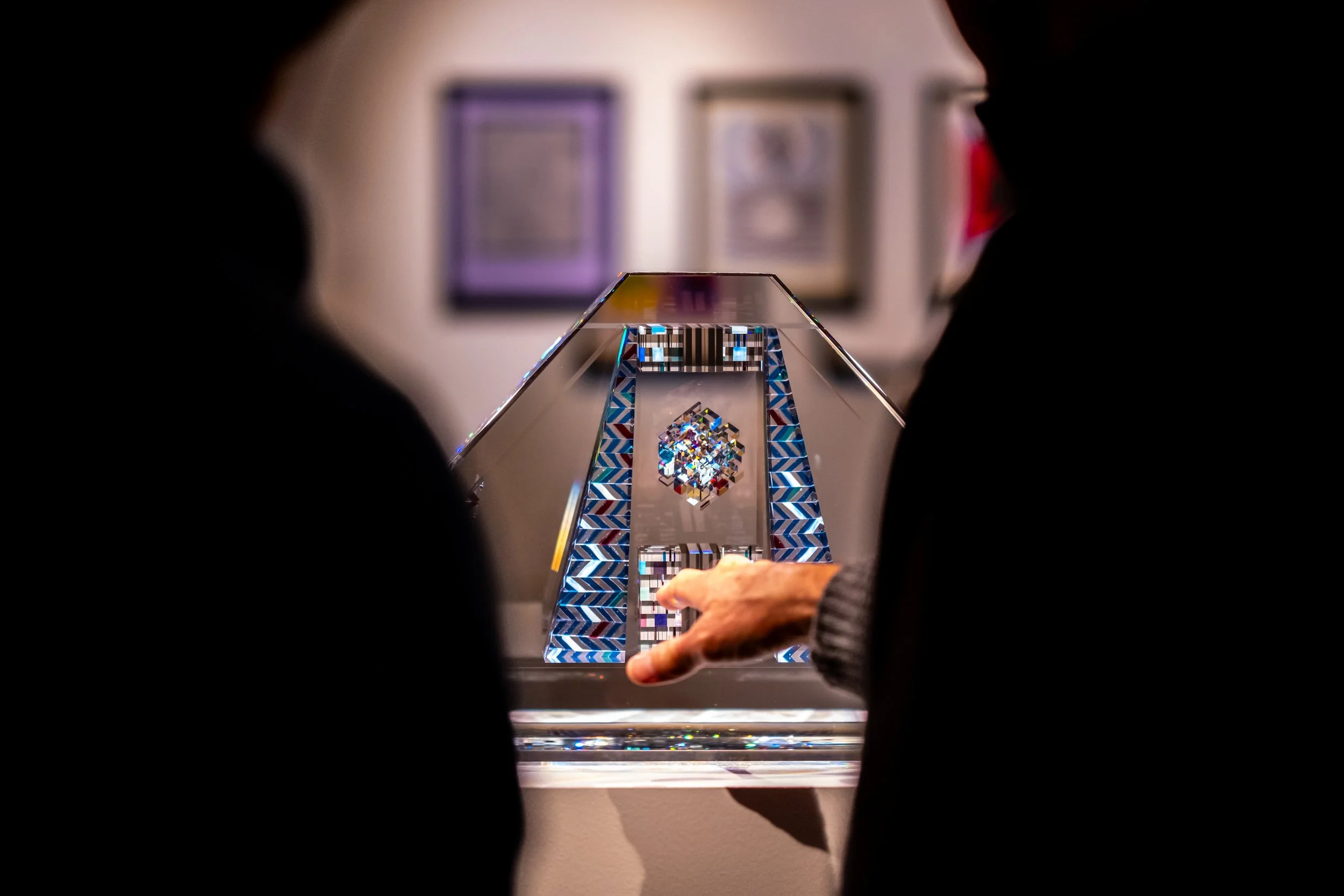 A person looks at a Tim Burton statue with a pyramid shape, featuring colorful patterns and a reflective surface, displayed in an art gallery.