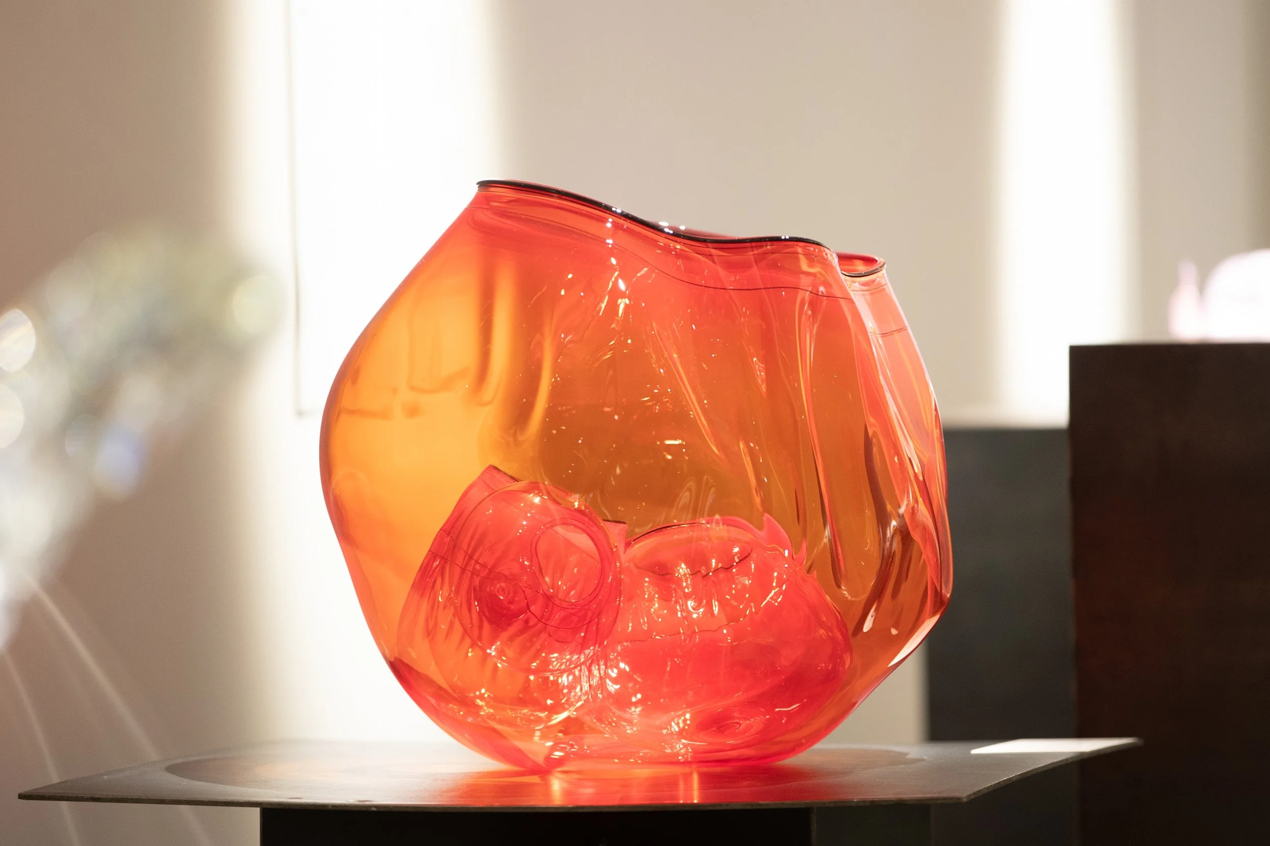 A close-up of a red glass sculpture with smooth, flowing curves, placed on a dark surface in a well-lit room.