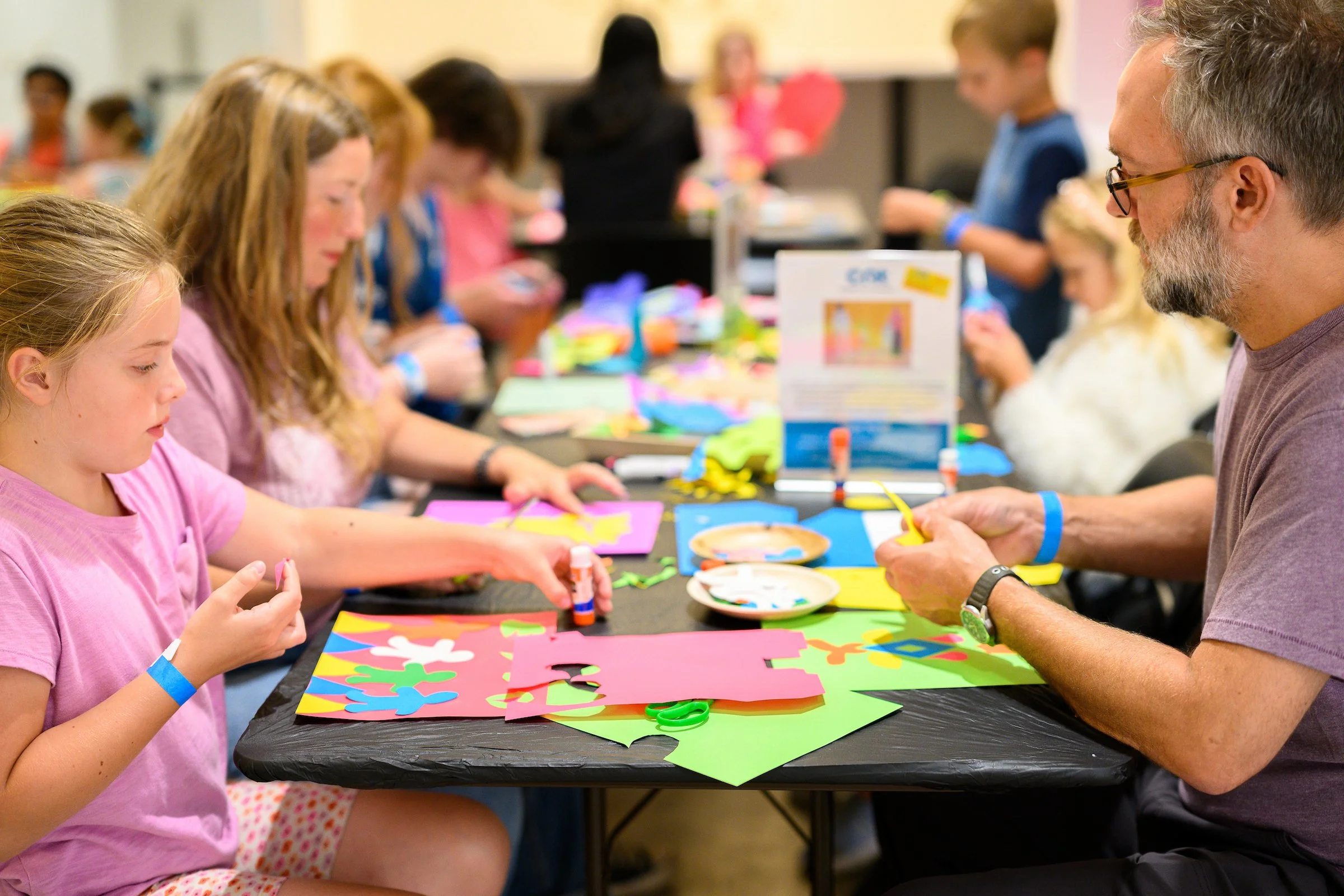 Adults and children seated at a table making collages of colorful neon paper.